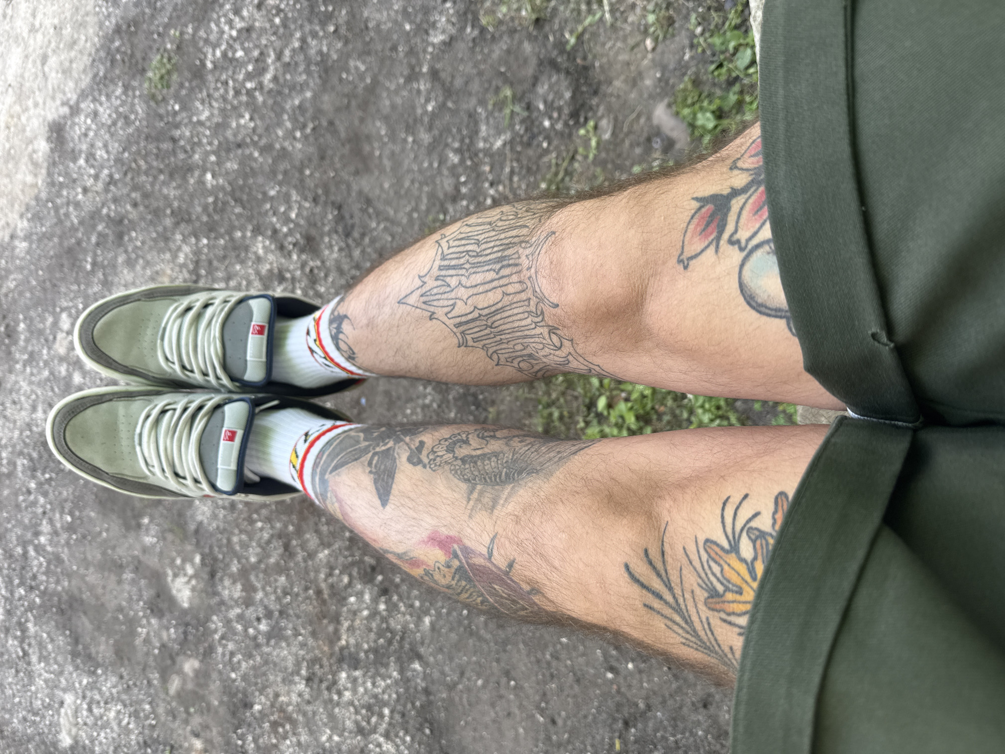 The image shows a first-person perspective of a person's legs and feet, sitting on a ground surface that appears to be gravel or dirt. The person is wearing olive green shorts, white socks with red and yellow stripes, and light-colored sneakers with gray accents and white laces. Their legs are heavily tattooed with various designs, including floral patterns, script, and other imagery. There is a notable red mark on the upper left thigh, possibly a bruise or ink. The background is out of focus and suggests an outdoor environment with some sparse green vegetation. The image was taken in Pleven, Bulgaria.