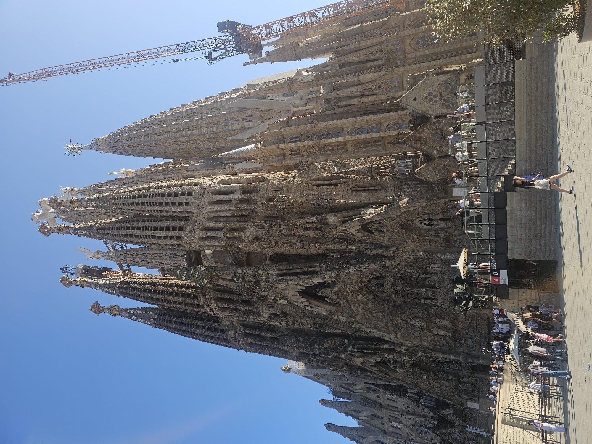 The image, captured in Barcelona, Spain, displays a section of the Sagrada Familia under a clear blue sky, indicating fair weather and likely daytime. The iconic basilica's intricate, organic architecture dominates the frame, with its towering spires and detailed facade. Construction cranes are visible in the background, suggesting ongoing work on the landmark. In the foreground and midground, people are gathered, some standing and observing the basilica, others walking along the paved area adjacent to it. One woman in the foreground is captured mid-stride, looking towards the building. The overall scene conveys a sense of tourism and admiration for the architectural marvel, with a relaxed atmosphere suggested by the casual attire of the visitors. The sun appears to be high in the sky, casting subtle shadows. No visible text is present in the image.