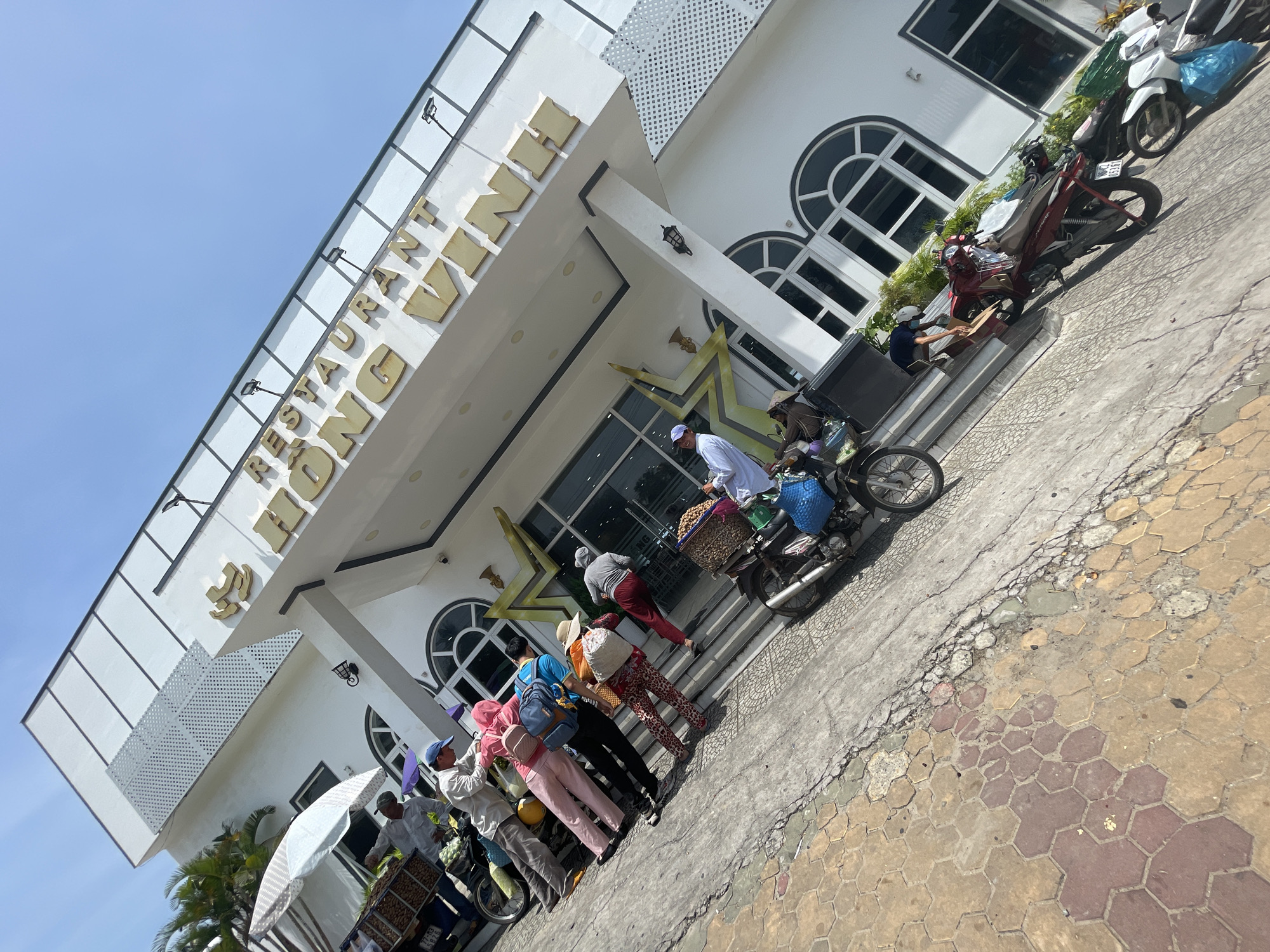 In Phu Thuy Ward, Vietnam, a group of individuals are gathered outside a white building with gold lettering that reads "RESTAURANT HONG VINH." The building features large arched windows and a prominent, star-shaped gold emblem above the entrance. Several motorcycles are parked in front of the building. Two individuals are near a motorcycle laden with baskets and what appears to be produce, possibly selling or unloading goods. Another group of three people, dressed in casual clothing, are standing near a motorcycle, with one person appearing to hand something to another. Further down the steps, a person in a white outfit is interacting with a motorcycle, seemingly adjusting or loading items. The ground is a mix of concrete and hexagonal paving stones. The scene suggests a busy moment at the restaurant, potentially involving delivery or a gathering of people with their vehicles and goods.