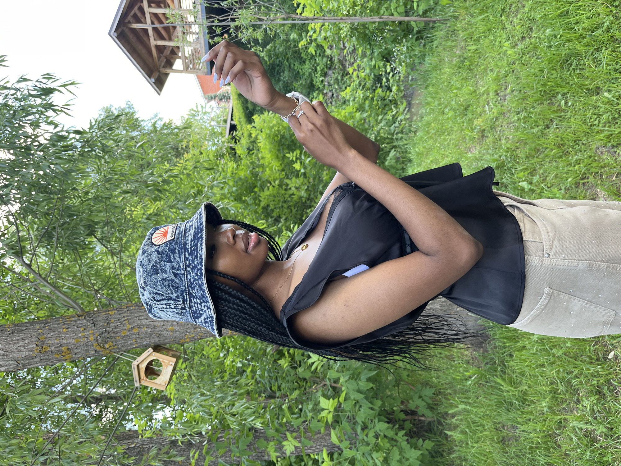 A young Black woman wearing a denim bucket hat, a black top with ruffled sleeves, and beige pants adorned with rhinestones stands in a garden with lush green trees and grass. She is looking down at her hands, which are adorned with rings and bracelets. In the background, a wooden birdhouse hangs from a tree, and a wooden structure with a roof is visible on a hill. The scene appears to be a serene outdoor setting. There is no indication that this scene is in Kazan, Russia.