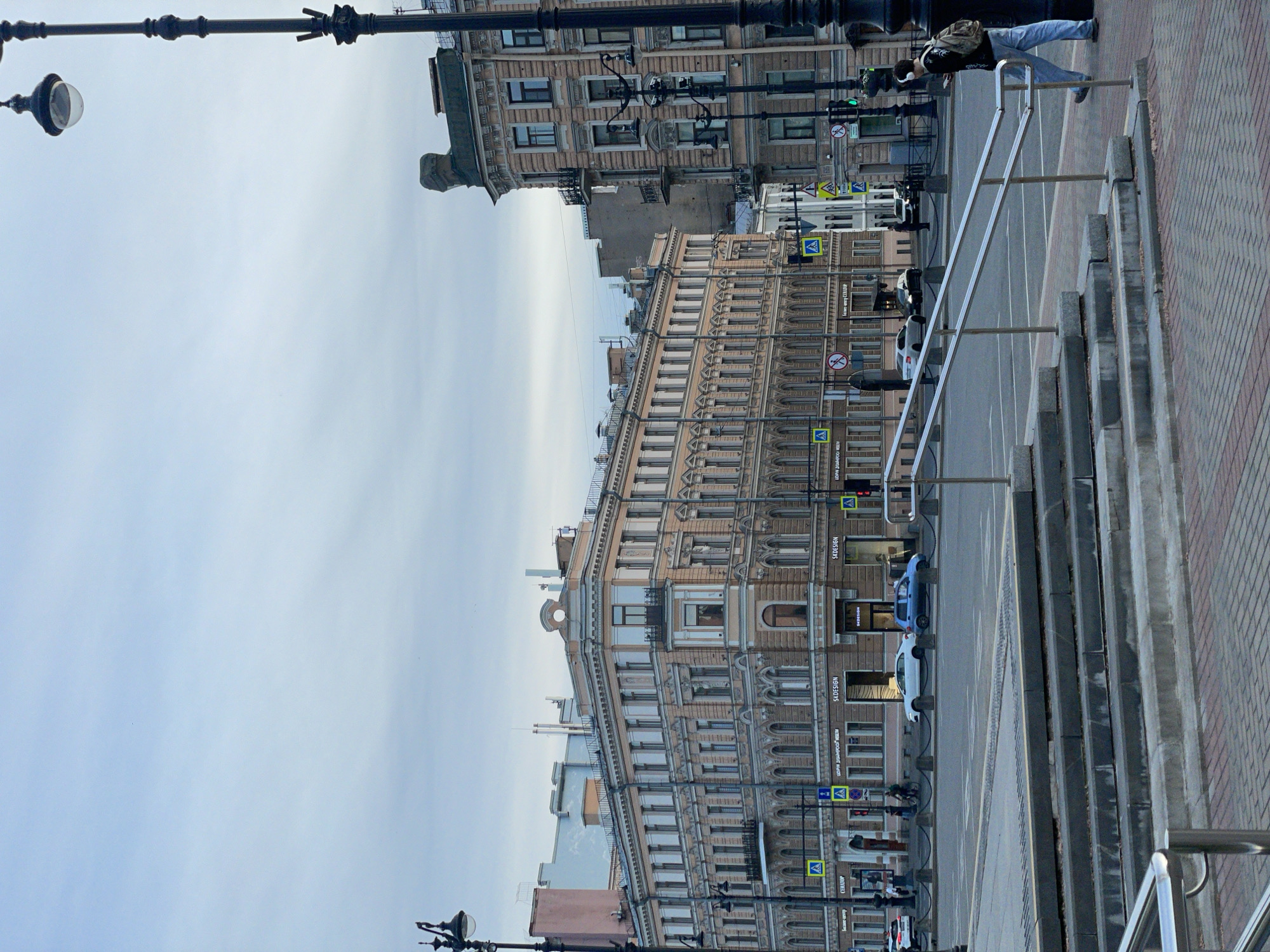 This is an overcast daylight street scene captured in Saint Petersburg, Russia, depicting a wide urban intersection bordered by historic architecture.
The prominent building on the left and center is a five-to-six-story structure with a pale peach-colored facade, featuring a classical architectural style with numerous tall windows, decorative pilasters, cornices, and sculptural elements in niches on the upper floors. The ground floor houses several businesses, including "SKODESIGN" and "ЦЕНТР ДИЗАЙНЕРСКОЙ МЕБЕЛИ" (Center for Designer Furniture) in Cyrillic, along with "СЕНАТОР" (Senator). To the right, another multi-story building, similar in architectural style but with a slightly darker, more weathered appearance, features a distinctive, ornate, dark-colored cap or chimney structure on its top-right corner.
In the immediate foreground, a section of brick-patterned sidewalk leads to a set of grey concrete steps with a modern silver metal handrail. Beyond these steps, a wide asphalt road or square stretches across the middle ground, separated from the sidewalk by a line of low black bollards connected by chains. Faint white road markings are visible on the asphalt surface.
A white sedan is parked or stopped on the street in front of the main building, followed by a blue compact car. Other vehicles are faintly visible further down the street. On the right side of the foreground, a male figure is walking leftward, wearing light blue jeans, a dark jacket, a light-colored backpack, and white over-ear headphones. He holds and looks at a smartphone in his left hand, mid-stride with his right foot raised. Several smaller figures are visible in the background, walking on the distant sidewalks.
The scene is illuminated by an overcast sky, casting a soft, diffuse light. Multiple tall, ornate black streetlights with spherical lamps are positioned throughout the scene, including one partially cropped on the far left and a dominant pole on the far right. Blue pedestrian crossing signs and yellow 'A' signs are visible on street poles and affixed to building facades, alongside a red traffic light on the right.