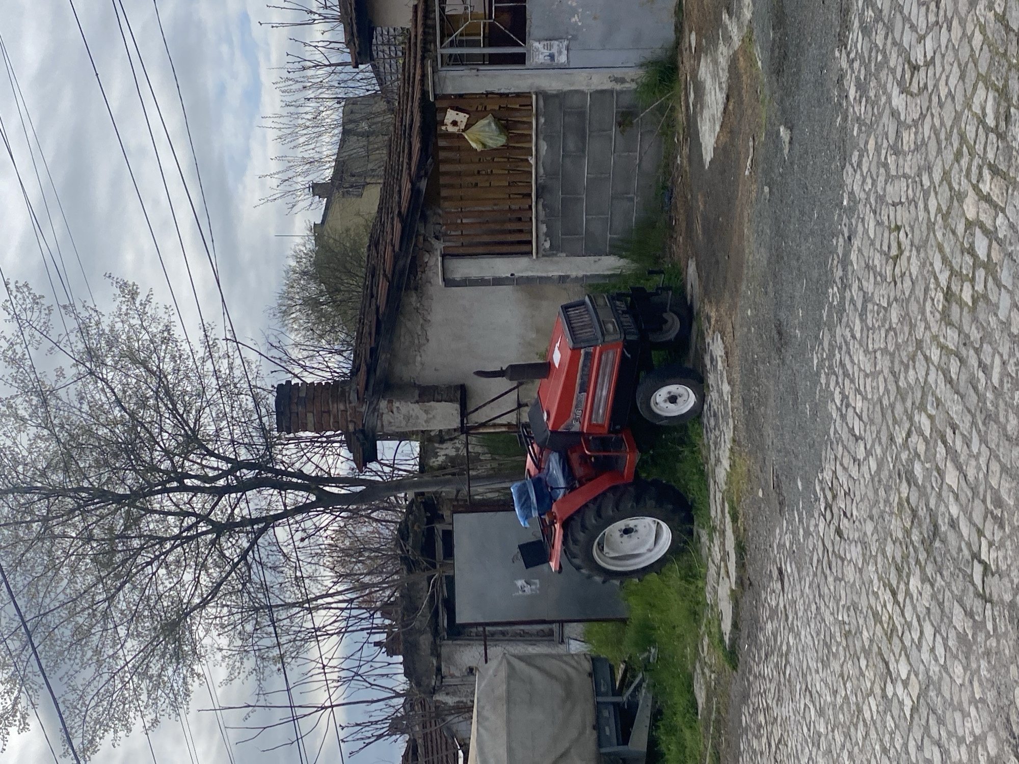 The image depicts a rural scene, likely in a neglected or transitioning urban area. The primary object is a small, red tractor parked adjacent to a dilapidated building with crumbling walls and a partially collapsed roof. The building exhibits a mix of construction materials, including concrete blocks, wood, and brick. Adjacent to the tractor and building, there is a paved area with uneven stone blocks. A bare tree with budding leaves dominates the left side of the frame, partially obscuring the view. Overhead, power lines traverse the scene against a cloudy sky. The overall impression is one of decay and stagnation amidst the beginnings of spring. There are no visible people, subjects, actions, or interactions. It is impossible to confirm the specific location in Burgas, Bulgaria, based on the image alone.