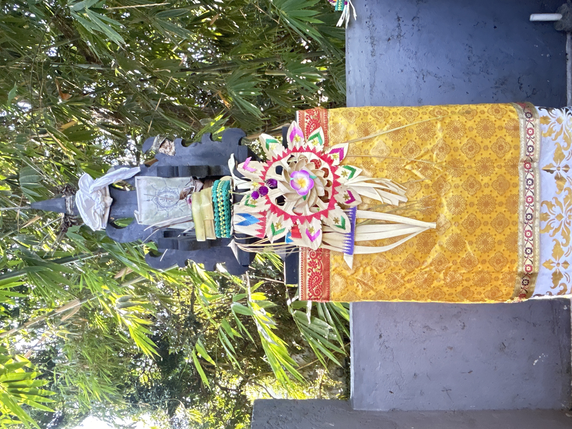 This image depicts a Balinese offering, known as a Canang Sari, placed on a stone structure, likely a shrine or altar, in North Kuta, Indonesia. The offering is adorned with vibrant yellow fabric, intricately patterned with floral motifs, and features decorative elements crafted from palm leaves, including a flower-like centerpiece in pink and purple. A woven green band wraps around a dark, decorative wooden stand, which holds small offerings or religious items. The background is dominated by lush green bamboo foliage, bathed in bright sunlight, suggesting an outdoor setting. The stone structure appears weathered, with a textured grey surface. The overall scene conveys a sense of cultural and religious significance within a natural environment.
