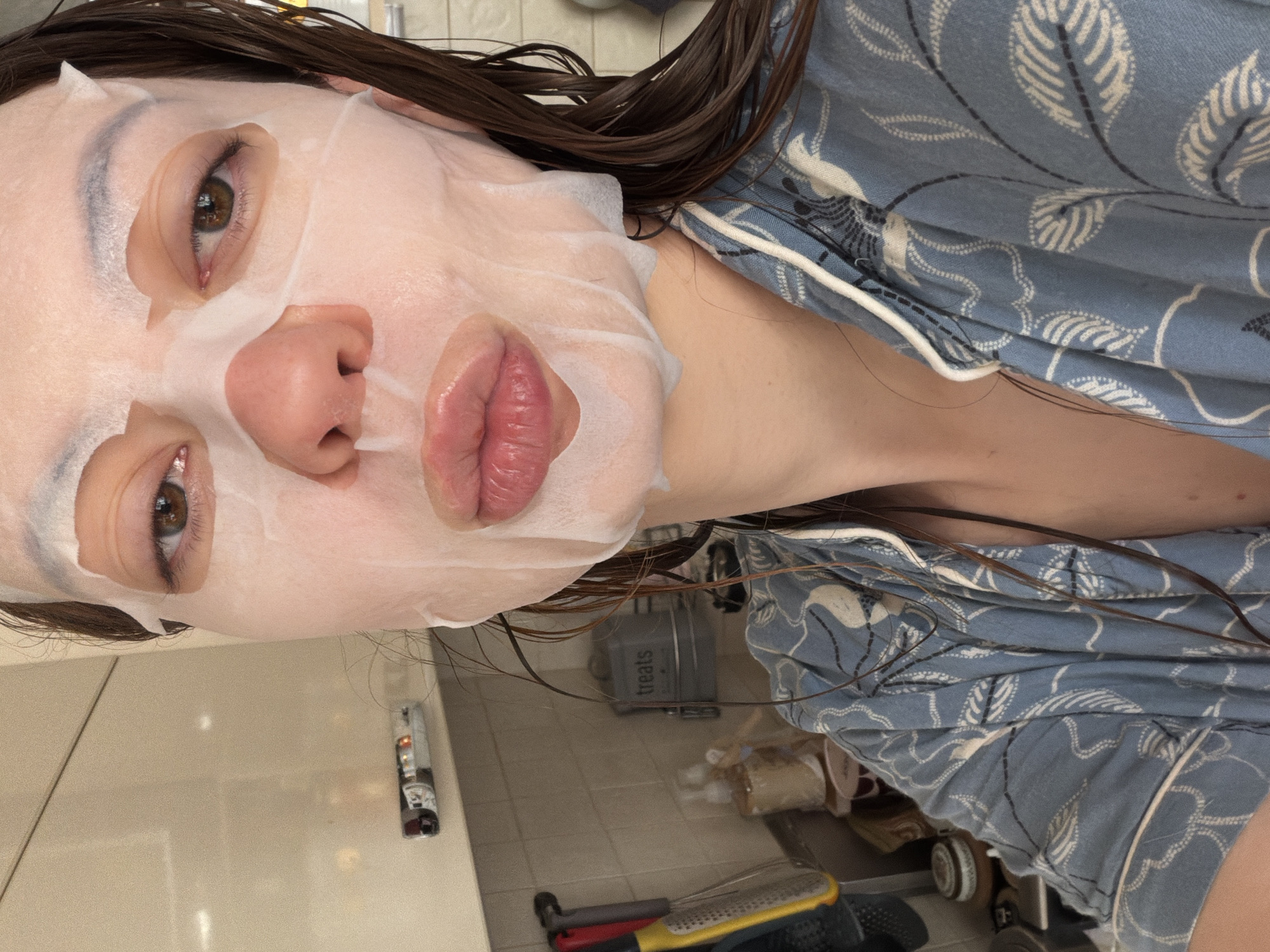 A woman is wearing a sheet mask on her face. She has light skin, brown hair, and brown eyes. She is wearing a blue patterned pajama top with white piping. Her hair appears wet. The background includes a tiled wall, what appears to be a counter with various objects including kitchen utensils and containers. The container labeled "treats" might be a decorative box. The location is not discernable from the image content. There is no evidence indicating it is located in Khimki, Russia.