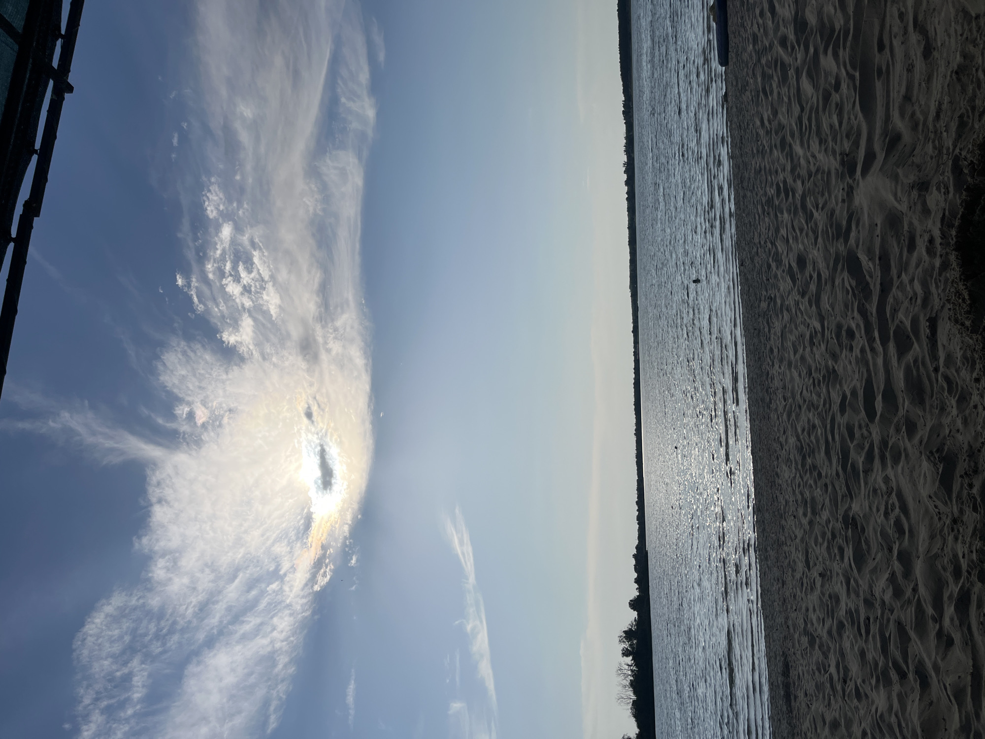 This image depicts a scenic view of a beach and the sky. The foreground on the right shows a sandy beach with rippled patterns, characteristic of windblown sand. To the left of the beach is a body of water, likely a lake or sea, with sunlight glinting off its surface, creating a shimmering effect. The water appears calm with small ripples.
The upper left portion of the image is dominated by a clear blue sky with wispy white clouds. The sun is partially obscured by these clouds, creating a halo effect and casting a bright glow. The clouds are elongated and streaky, suggesting a light breeze.
Along the horizon, a thin line of dark trees marks the opposite shore of the body of water. The overall scene suggests a peaceful outdoor setting during the daytime, possibly late afternoon given the angle of the sun and the lighting. There is no discernible activity or event taking place, and no people or specific objects are prominently featured, apart from the natural elements of sand, water, sky, and trees. The mood is serene and tranquil, evoking a sense of natural beauty and calm. The location is identified as Yelyzavetivka, Ukraine. No text is visible in the image.