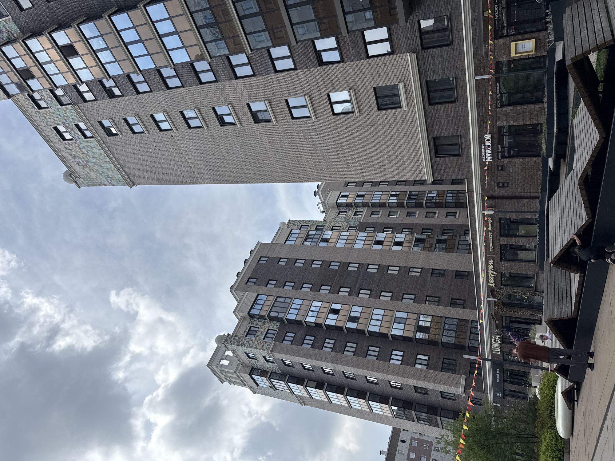 This is a medium shot, eye-level, outdoor, daytime photograph of two modern apartment buildings in Krasnoyarsk, Russia. The building on the right is taller and closer, with a light brick facade and darker brick accents around the windows. It has numerous balconies with dark tinted glass. The building on the left is shorter and slightly further away, with a darker brick facade and similar window treatments. Both buildings feature decorative tiled sections near their roofs. A street with several storefronts is visible at the base of the buildings. Signs indicate businesses such as "LUNGA" and "АБСОЛЮТНО МУЖСКОЙ." Small, colorful triangular flags are strung across the storefronts. In the foreground, a man wearing a brown shirt and grey pants stands in an open plaza area. He is facing away from the camera and appears to be holding a thin, dark pole. A child in a white shirt is partially visible behind him. To the right, another person, a child, is interacting with a wooden structure in the plaza. The plaza is paved and features several wooden, tiered seating or lounging areas. The sky is overcast with visible clouds.