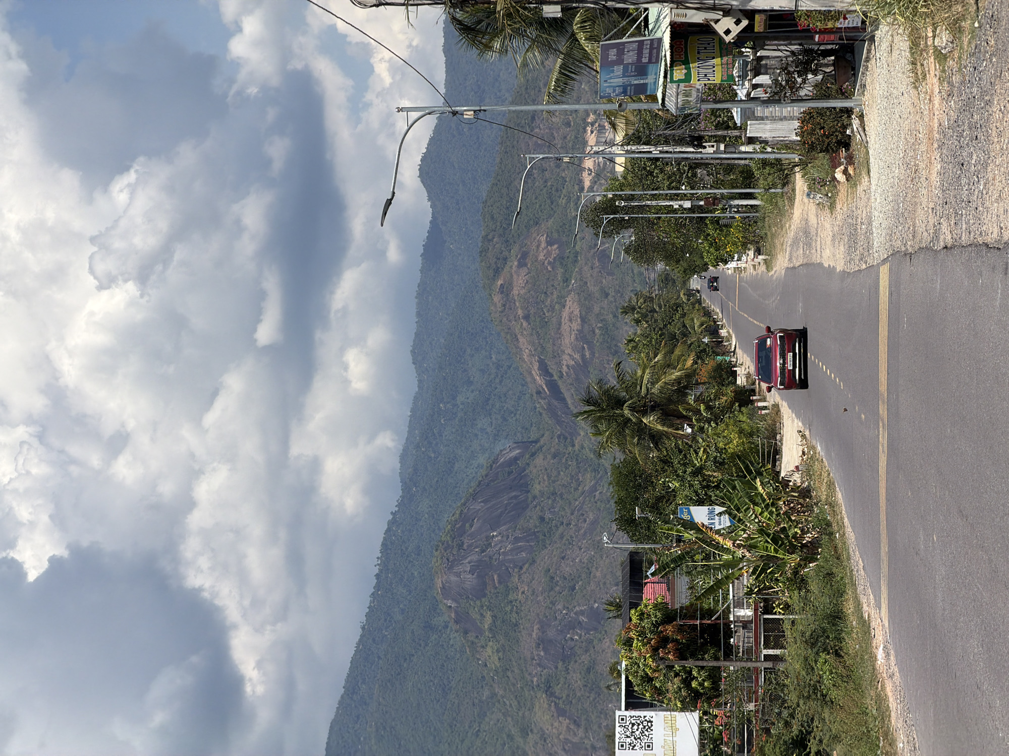 An outdoor view in Diên Thọ, Vietnam, features a road stretching into the distance with a red car driving on it. The roadside is flanked by vegetation. A mountain covered in trees looms in the background, contrasting with a cloudy sky. Overhead, streetlights line the road. Signage and buildings are visible along the roadside.