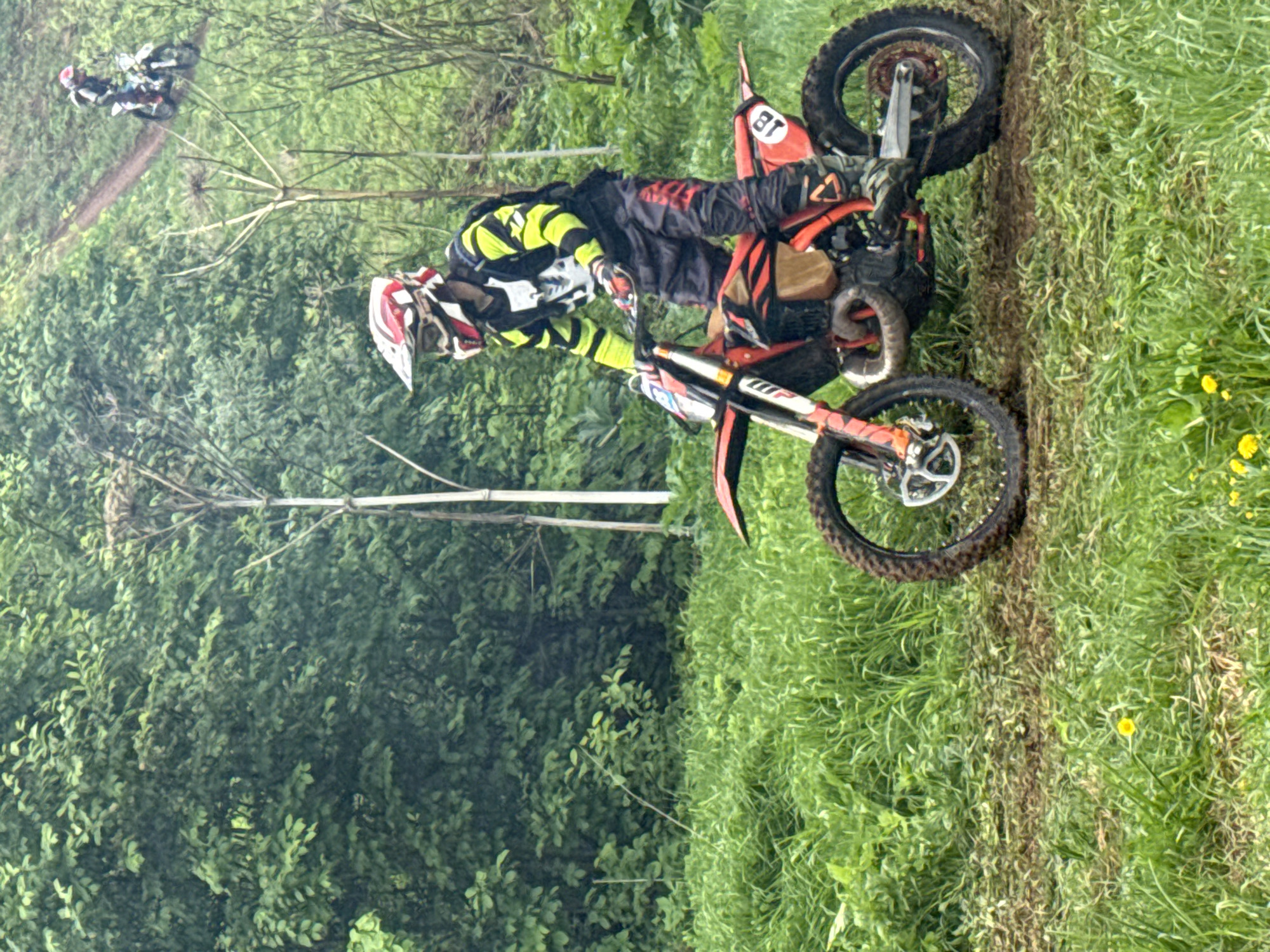A dirt biker wearing a helmet and riding gear is maneuvering a motorcycle on a grassy track with a path of disturbed earth, with another biker on a motorcycle visible in the background. The setting appears to be Чёрная, Russia, judging by the natural environment visible in the image. There is dense foliage and trees along one side of the track, with wildflowers and grasses visible on the other. The motorcycle has the number "81" on it.