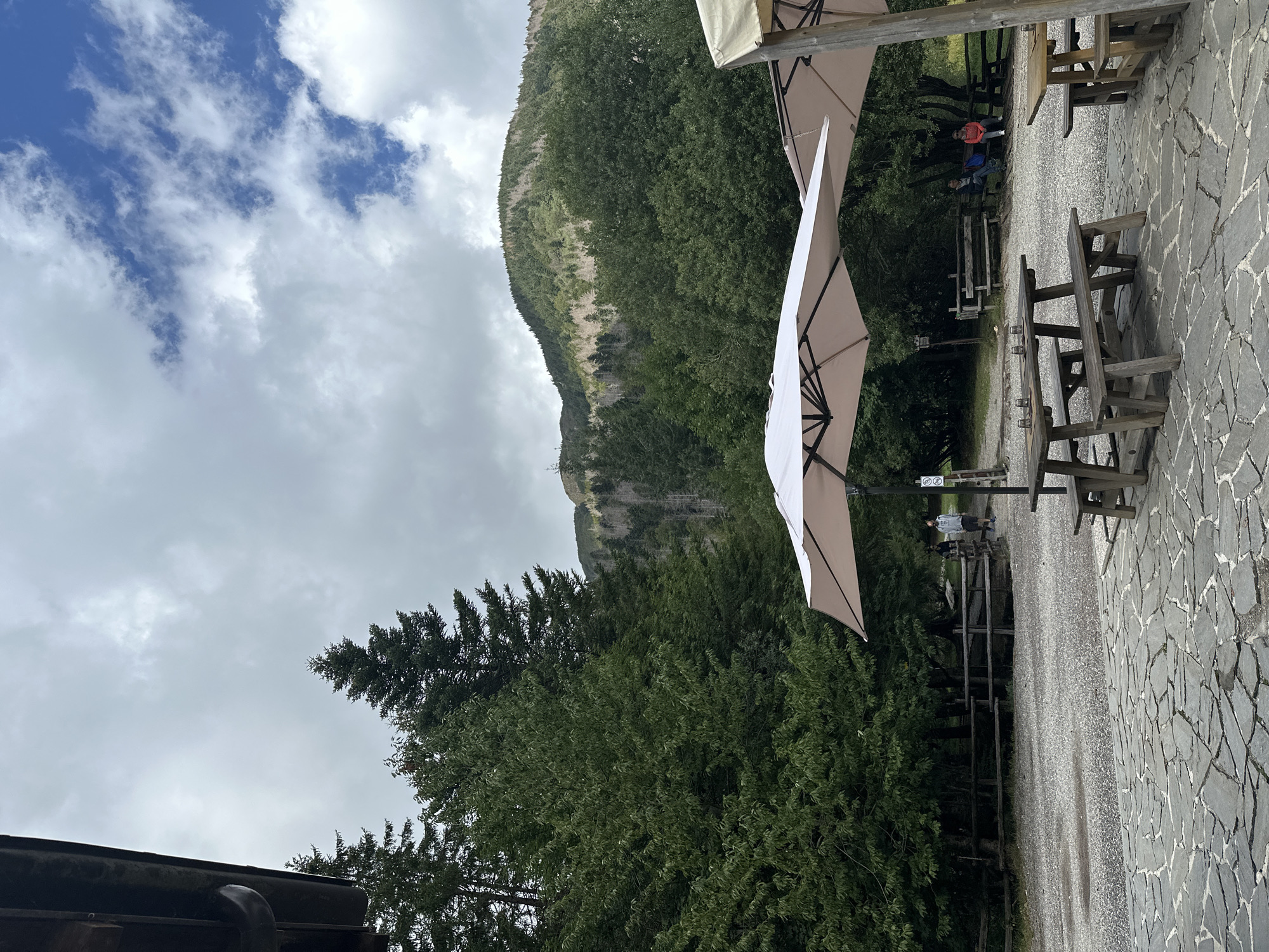 This image depicts an outdoor seating area with wooden picnic tables and benches under large, light-colored umbrellas. The ground is paved with irregular stone slabs in the foreground and gravel in the background. A wooden fence separates the seating area from a wooded area with tall evergreen trees. In the distance, a green, tree-covered mountain rises under a cloudy sky. Two people are visible walking in the background, and two people are sitting on a bench in the middle ground. The scene appears to be in a natural setting, likely a park or a restaurant with an outdoor patio, and could potentially be located in or near Corniglio, Italy, given the mountainous and forested environment.