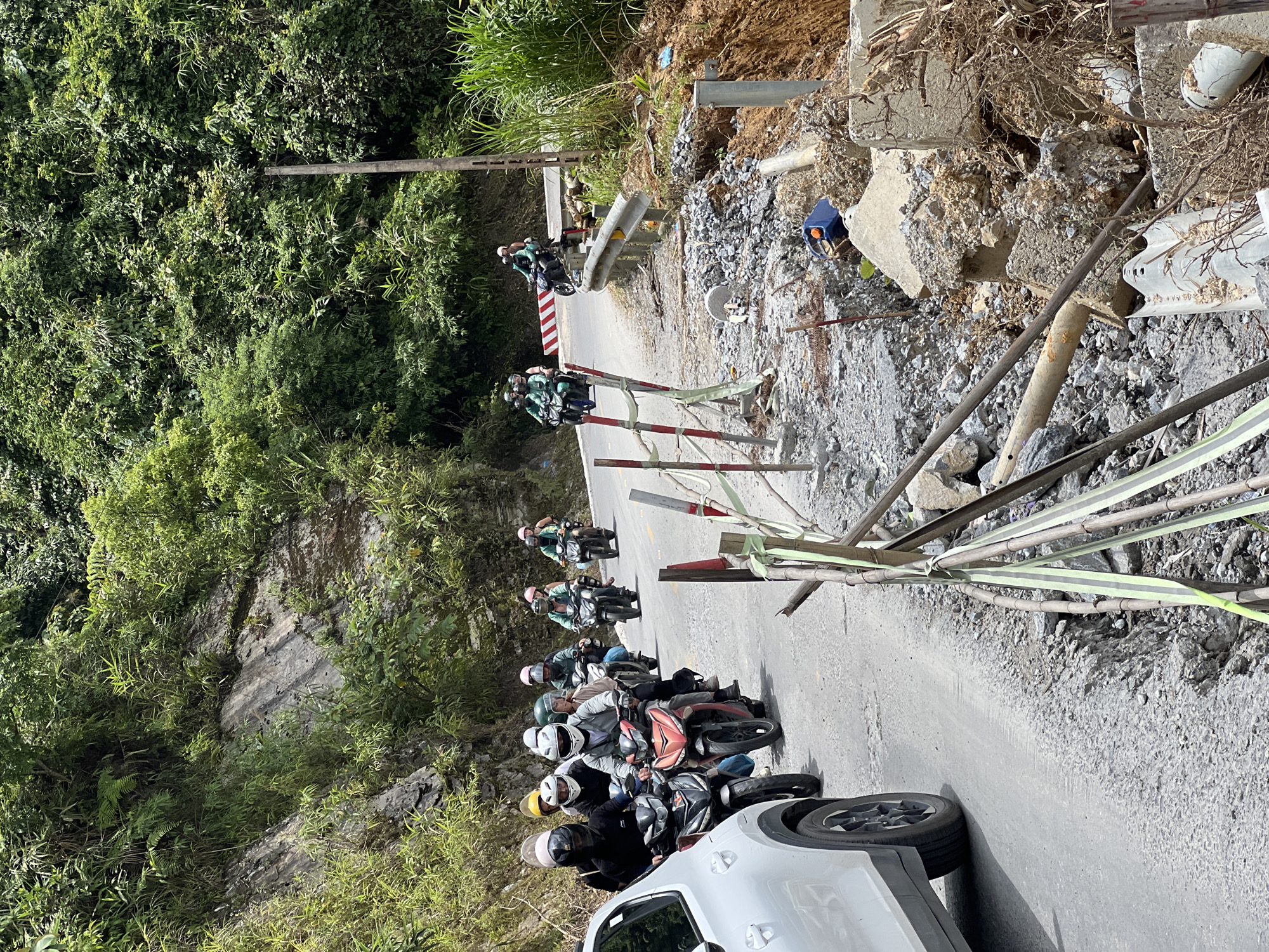 On a paved mountain road in Quản Bạ Commune, Vietnam, a procession of motorcyclists navigates past a section of the road that has suffered a landslide. A white car is partially visible on the left, traveling in the same direction as the motorcycles. The landslide has caused debris, including rocks, soil, and broken concrete, to spill onto the road and down the embankment. Temporary barriers constructed from bamboo and caution tape are in place to mark the damaged area. The surrounding environment is lush with dense green vegetation covering the steep hillsides. Several motorcyclists, all wearing helmets, are visible in the frame, with some riding in pairs. The road surface appears worn and shows signs of damage beyond the landslide area. The sky is bright and appears to be daytime.