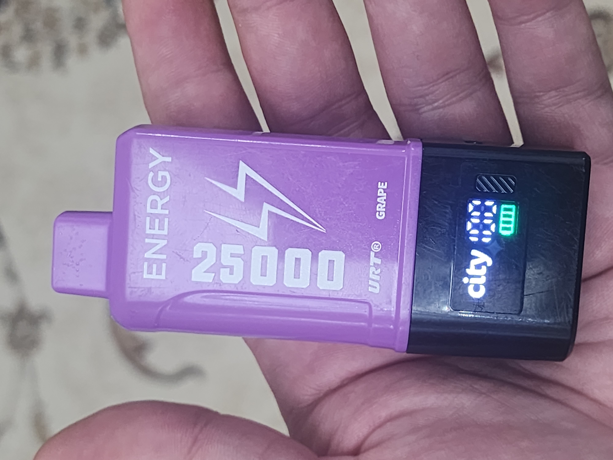 The image shows a person's hand holding a purple and black electronic smoking device, commonly known as a vape. The device has text printed on it, including "ENERGY," "25000," "URT®," and "GRAPE." A small digital screen on the device displays the word "city" and a battery indicator with a green bar. The background appears to be a patterned surface. There is no visible indication of the city of Gudermes, Russia.