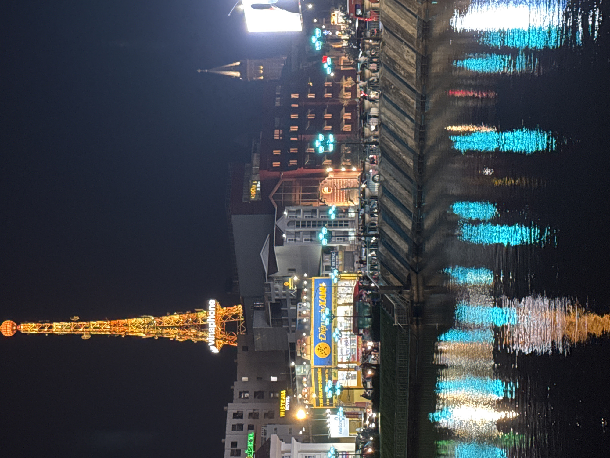 The image depicts a vibrant nighttime scene in Da Lat, Vietnam, likely along a river or waterfront. The orientation of the image is rotated 90 degrees counter-clockwise. The main focus is a brightly lit tower adorned with numerous lights, showcasing the "Vinaphone" logo. Below and to the right of the tower, a cluster of buildings is illuminated with a variety of colorful lights, including neon signs with Vietnamese text like "WISTERIA HOTEL," "Điện máy XAN," and "VÔ HƯƠNG."
Reflections of the lights create a dazzling display on the dark water, adding to the lively atmosphere. The scene appears to be a bustling commercial or entertainment area, with hints of people visible on balconies and near the shops. The time of day is clearly night, as indicated by the dark sky and the prominent artificial lighting. The weather appears to be clear, allowing for the vivid reflections on the water. The overall impression is one of a lively and visually dynamic urban environment at night.