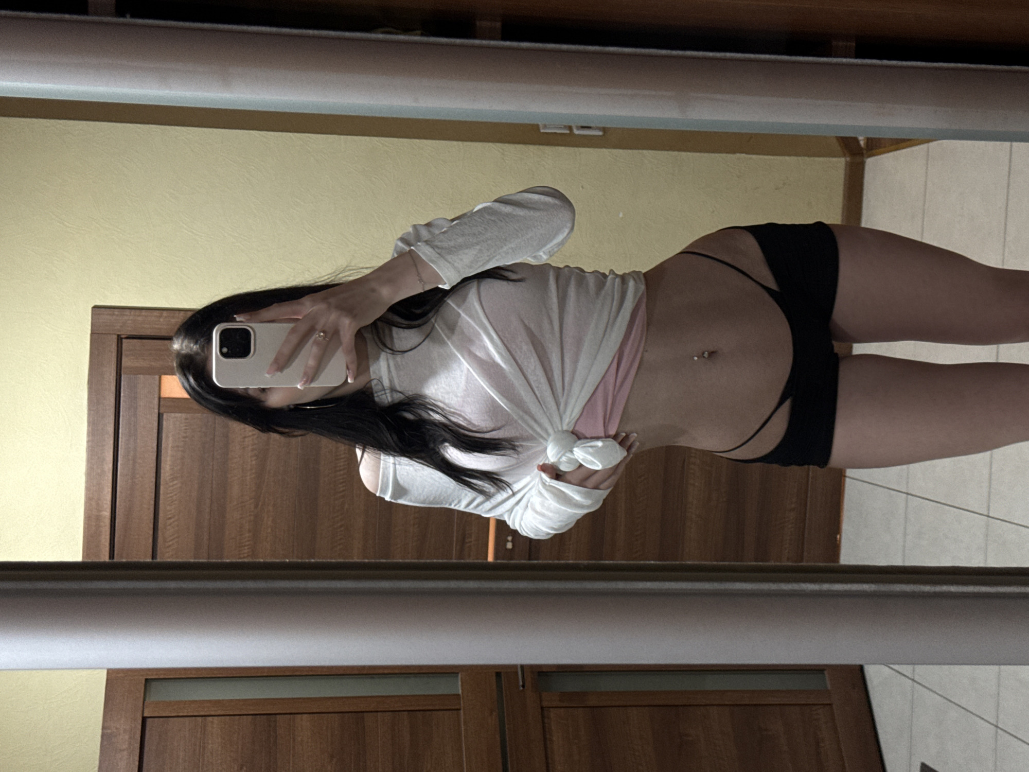 A young woman with dark hair takes a mirror selfie in an indoor setting. She is wearing a sheer white off-the-shoulder long-sleeved shirt tied at the waist over a pink undergarment, and black shorts. Her navel is pierced. The background shows wooden doors and a yellow wall. The location is identified as the city of Khimki, Russia.