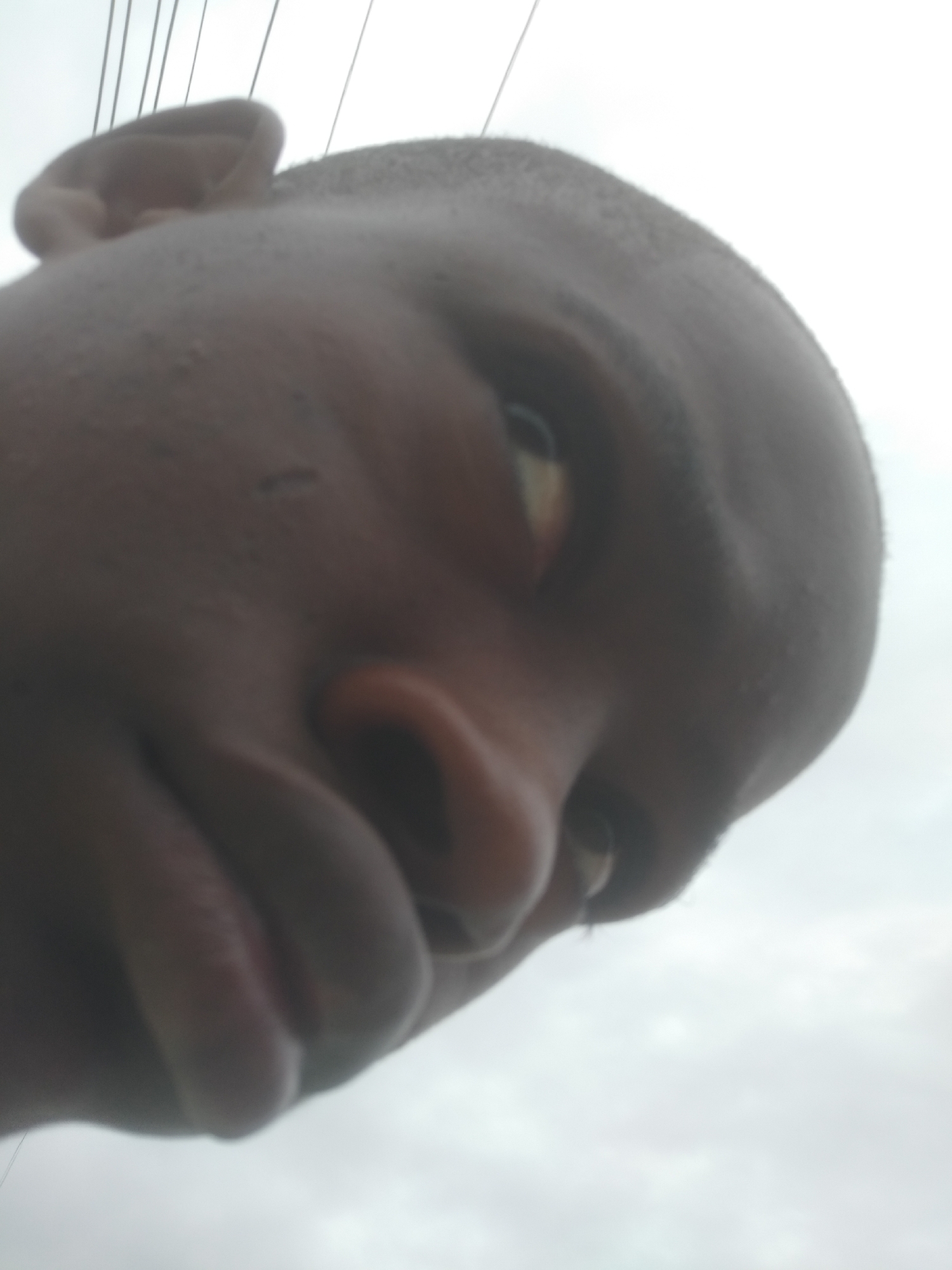 A close-up, low-angle shot shows a person's face, angled downwards toward the camera. The person has dark skin, a shaved head, and is looking slightly to the side with one eye more visible than the other. The person's nose and lips are prominent in the frame. Overhead, thin wires are visible against a hazy, overcast sky. The location appears to be outdoor. The city is unknown.