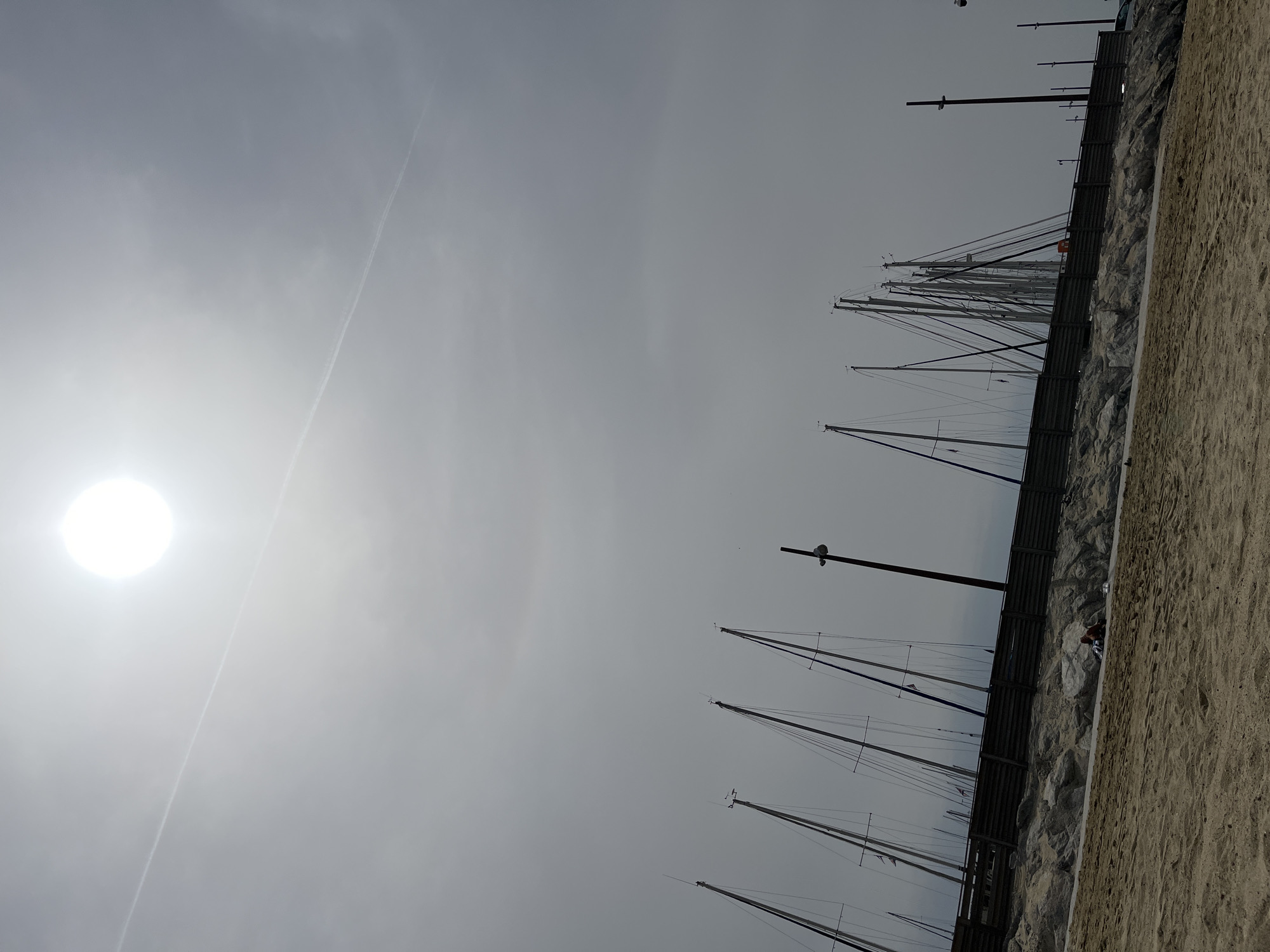 The image depicts a view of sailboats moored in Sainte-Maxime, France, viewed from a low angle. The sky is overcast, with the sun appearing as a bright, diffuse circle. There is a contrail visible in the sky. A line of sailboats with masts of varying heights runs diagonally across the frame. A fence runs alongside the masts. A beach area with sand is on the right.