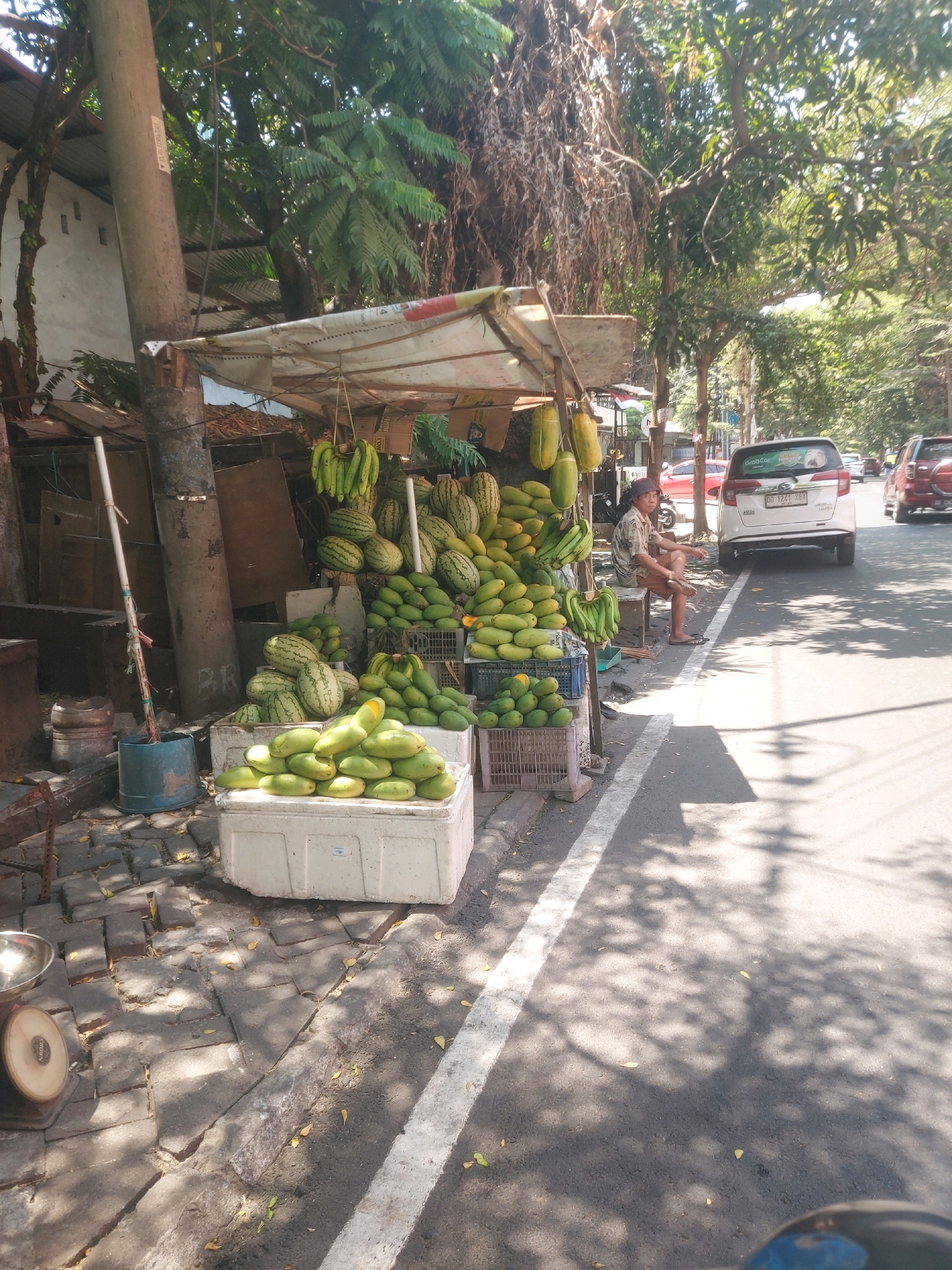 An outdoor market stall displaying a large assortment of fresh fruits is captured on a sunny day on the side of a paved road in Makassar, Indonesia. The stall is set up under a canopy, with various fruits arranged on the ground and on crates. Dominant fruits include watermelons, mangoes, and bunches of bananas. A person, dressed in a patterned shirt and shorts, sits on a bench behind the stall, facing the road. Several vehicles, including a white car with a "GrabCar" sticker and another red car, are visible on the road. The ground around the stall is paved with interlocking bricks, and scattered leaves are present. A weigh scale is positioned to the left of the stall. The environment suggests a typical street scene in a tropical urban setting, with shadows cast by trees and the overhead canopy.
