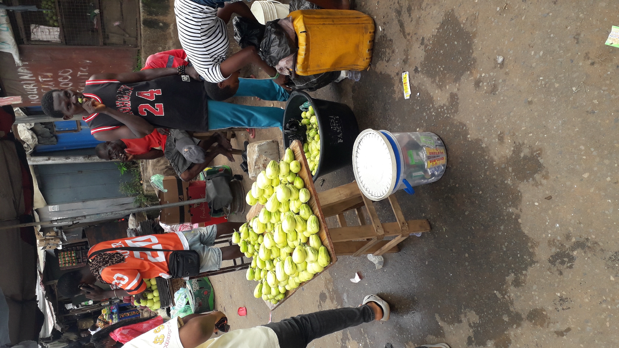 In Nkpor, Nigeria, a bustling outdoor market scene unfolds. A vendor, wearing an orange jersey with the number 20, stands on the left, engaged in a phone conversation. Nearby, a man in a red shirt and shorts sits, chewing on a green item. In the center, another man in a black basketball-style jersey with the number 24 also chews on a green item, looking towards the camera. A woman in a striped shirt is observed interacting with a large yellow container on the right. In the foreground, a wooden table displays a large mound of light green, striped vegetables, likely garden eggs, with more vegetables visible in a black bucket. A clear plastic bucket filled with various canned drinks sits on the wet, concrete ground. The environment appears to be a busy street market with weathered structures and a damp, textured ground.