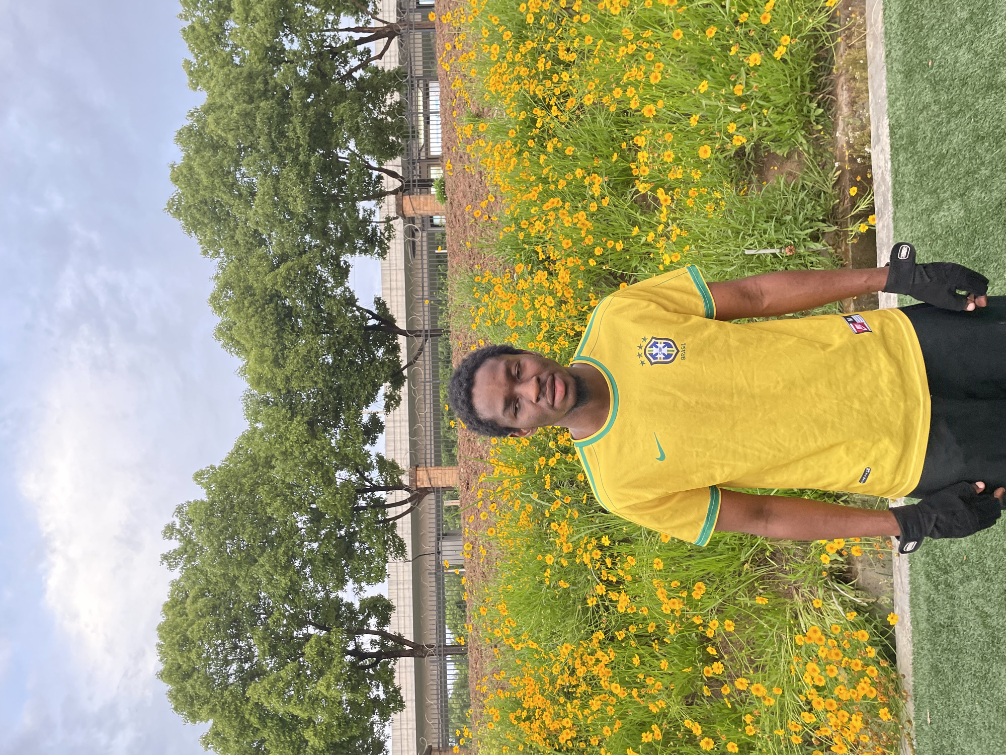 The image shows a man standing in a field of yellow flowers. He is wearing a yellow Brazil soccer jersey and black gloves. The flowers are growing in a field of green grass. Behind the man, there is a building with a metal fence, and trees are to his left. The location is Jiangjin, China. The sky is partly cloudy.