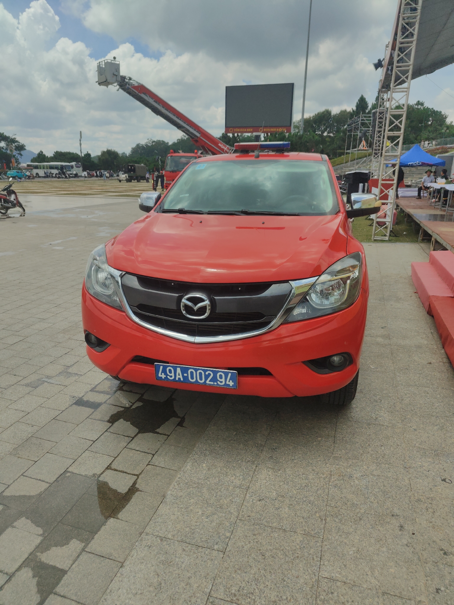 In Da Lat, Vietnam, on a cloudy day, a bright red Mazda BT-50 pickup truck, identified by its license plate "49A-002.94", is parked on a paved area. The truck has a blue and red light bar mounted on its roof. In the background, a large red fire truck with an extended ladder is visible. Further back, a large outdoor LED screen and a truss structure supporting stage lighting are present. People are seen in the distance, some near a blue tent and others seated on a red tiered platform. The ground is wet in patches, suggesting recent rain.