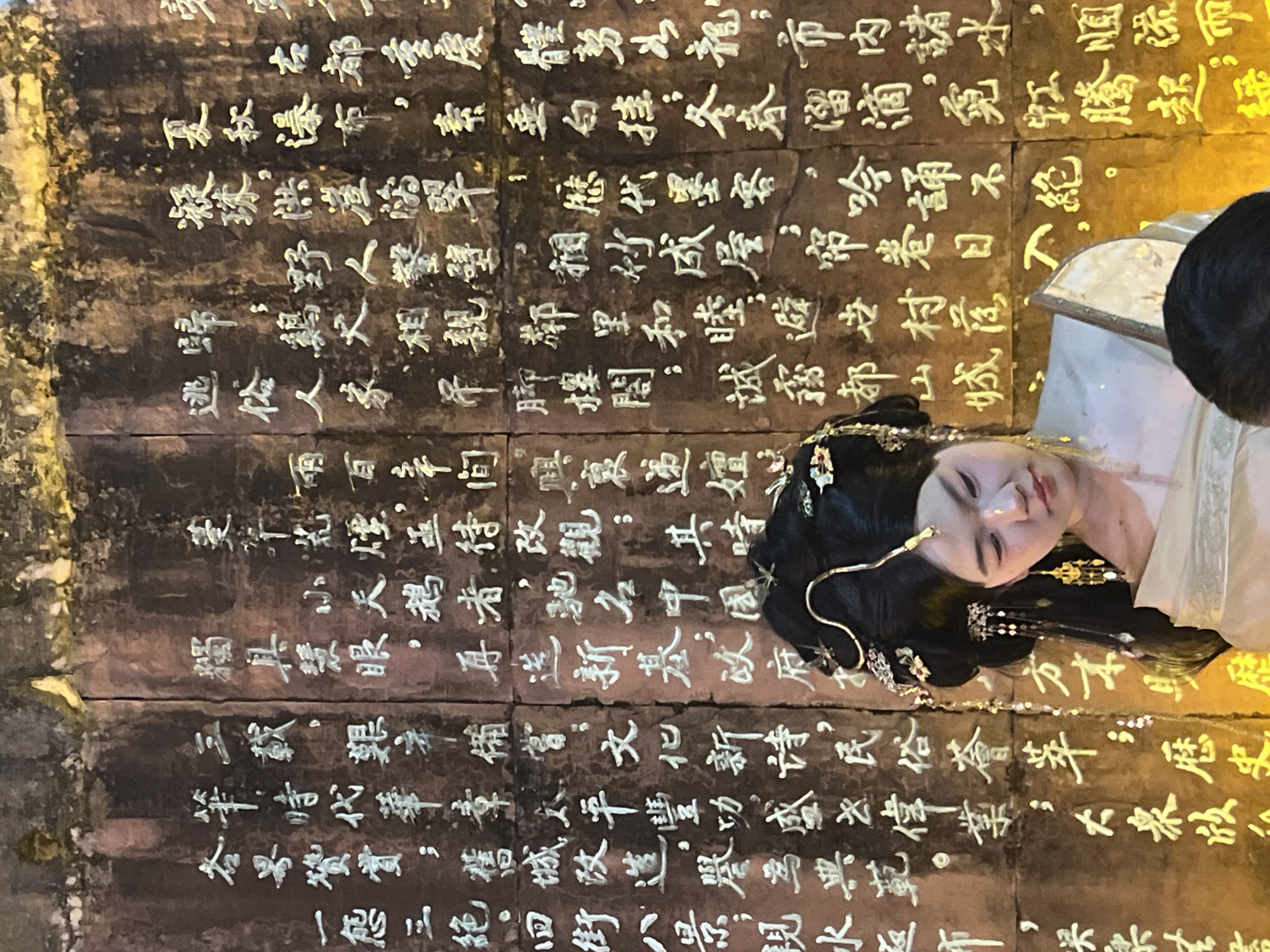 A woman in traditional Chinese attire poses in front of a wall of inscribed stone tablets. The text on the tablets is in traditional Chinese characters. The tablets are located in Yuzhong District, China. The text refers to historical landmarks and cultural achievements.