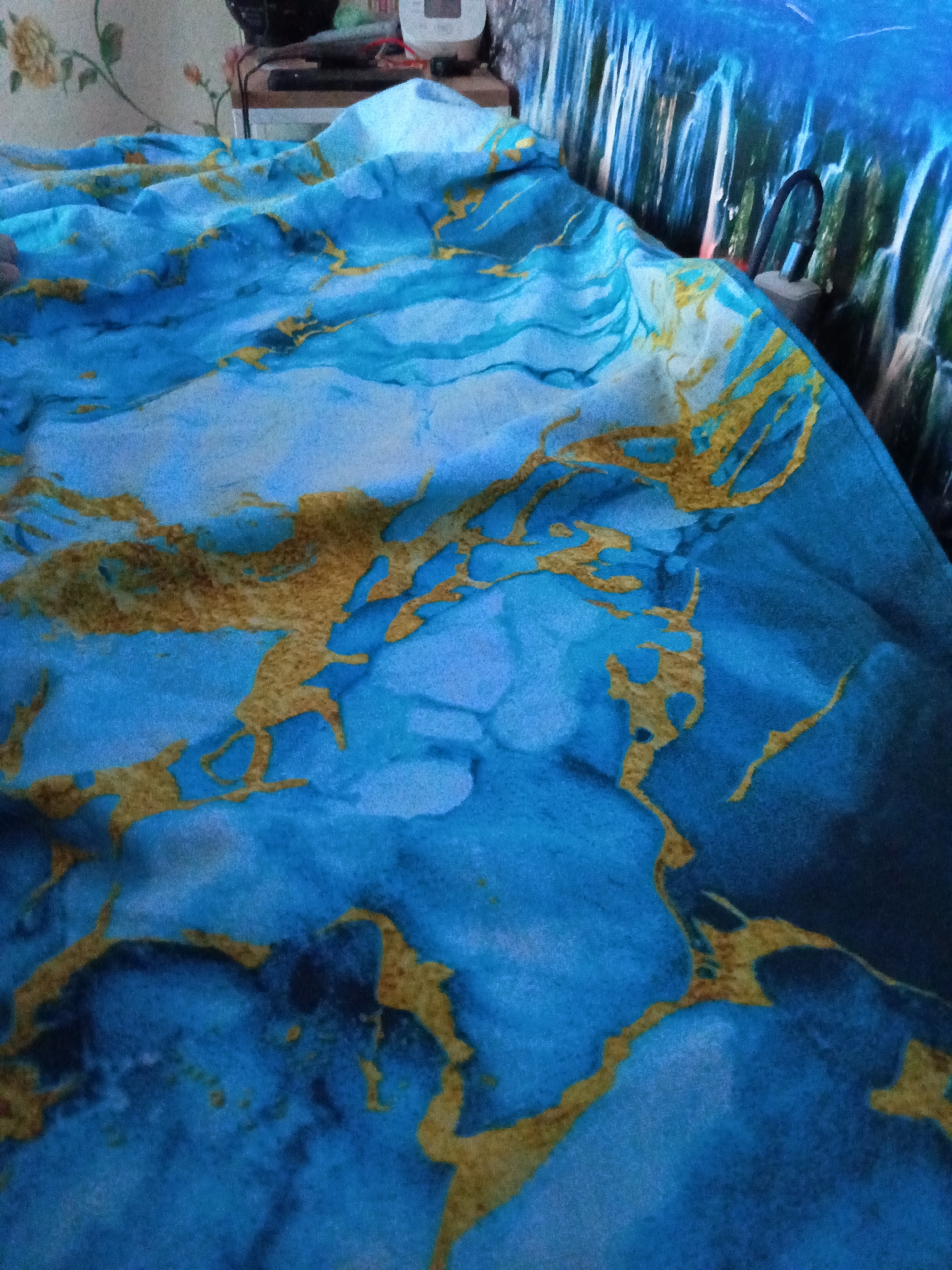 The image shows a close-up of a blue and gold patterned blanket or fabric. The pattern resembles marble or a swirling abstract design with veins of gold against a deep blue background. The fabric is wrinkled and appears to be draped over a surface, possibly a bed or couch. In the background, a wall with a floral pattern is partially visible, along with some objects on a shelf, including what appears to be an electronic device and some wires. The scene is indoors, and there is no indication of the city of Ust-Abakan, Russia, in the visual information provided.