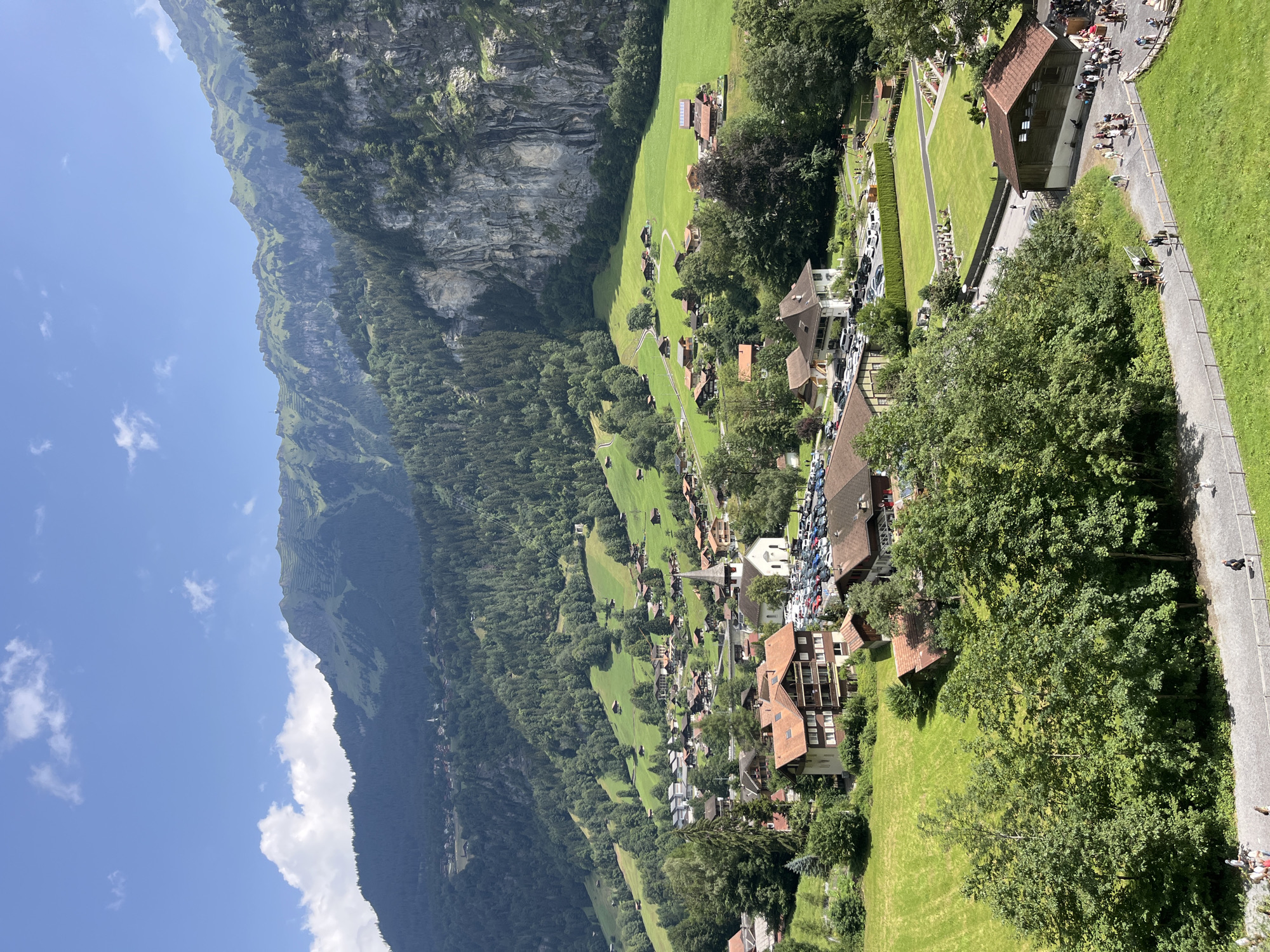The image depicts a panoramic view of the mountain village of Wengen, Switzerland. In the foreground, a paved pathway runs horizontally, bordered by lush green grass and trees. Several individuals are walking along the pathway. Beyond the pathway, more trees and grassy slopes lead up to a cluster of buildings that form the village.
The village architecture features traditional Swiss chalets with wooden balconies and steeply pitched, terracotta-colored roofs. A prominent white church with a tall steeple stands at the center of the village, surrounded by a parking lot filled with cars. Numerous houses and smaller buildings are scattered throughout the village, nestled amongst the trees and on the terraced hillsides.
In the background, the landscape rises dramatically into steep, rocky mountains covered in dense green forests. The slopes are further accentuated by terraced fields and the sheer rock faces of cliffs. The sky above is a clear blue, dotted with a few white, fluffy clouds. The overall scene conveys a serene and picturesque mountain village setting.