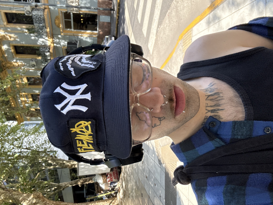 A person wearing a New York Yankees baseball cap with patches and glasses poses in front of a building. The cap has a white "NY" logo, a white skeleton hand patch, and a yellow patch with writing. The person has nose piercings and tattoos on their face and neck. They wear a black tank top and a blue plaid shirt. The background includes a building with decorative architectural details, a tree, and a crosswalk. The image is rotated sideways.