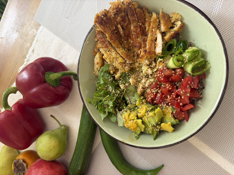 The image displays a detailed, elevated view of a prepared meal bowl situated on a table in Tel-Aviv, Israel. The circular, light green bowl, with a dark rim, occupies the lower central portion of the frame. Inside, the meal is meticulously arranged: the upper right quadrant contains numerous slices of golden-brown, breaded, and fried chicken, sprinkled with sesame seeds. The upper left quadrant holds a portion of mixed green salad leaves, also topped with sesame seeds. Below the greens, bright green, diced avocado pieces are visible, and further below, diced red tomatoes, also garnished with sesame seeds. The lower right features rolled slices of green cucumber. A base layer of what appears to be quinoa or another grain is visible beneath these ingredients. Adjacent to the meal bowl, on the left side of the table, various fresh produce items are arranged on a light-colored, textured fabric placemat. These include two large red bell peppers, two green pears, one red apple, one orange persimmon, one long green cucumber, and one long, thin green chili pepper. The table surface beneath the placemat is light-colored wood. Another light, subtly patterned placemat is visible on the right side of the bowl. No individuals are present in the scene. The overall impression is one of a freshly prepared, healthful meal.