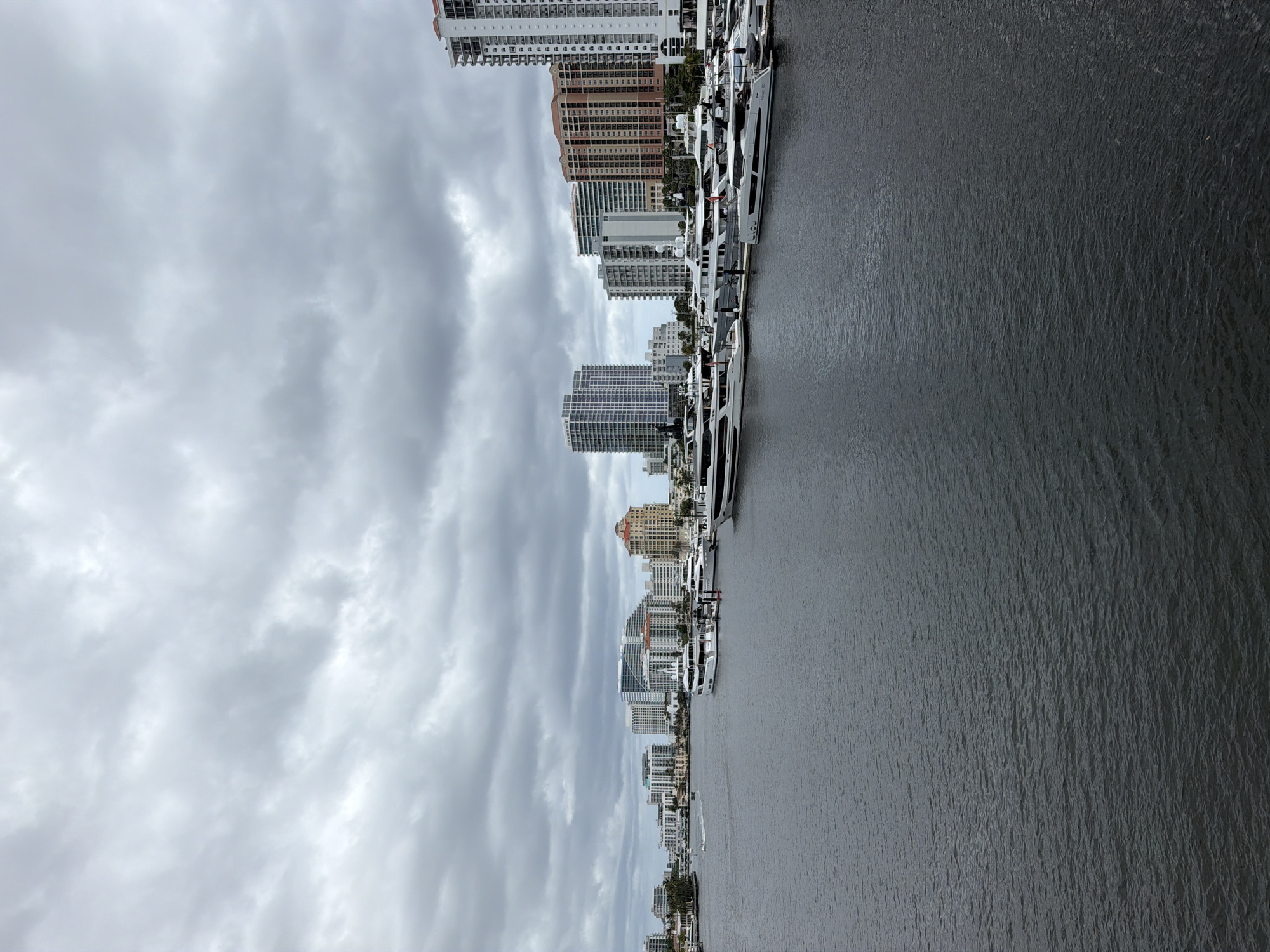 This photograph depicts a waterside scene featuring a row of modern buildings and numerous yachts docked along the water's edge. The architecture of the buildings is characterized by high-rise structures with varying designs and color palettes, indicating a relatively modern or redeveloped urban area. The sky is heavily overcast, suggesting a cloudy or potentially stormy day. The water appears dark and reflects the ambient light and surrounding structures. The yachts are neatly arranged, suggesting a marina or harbor area, and appear to be well-maintained. The photograph has been taken at an angle, causing the buildings to appear somewhat tilted.