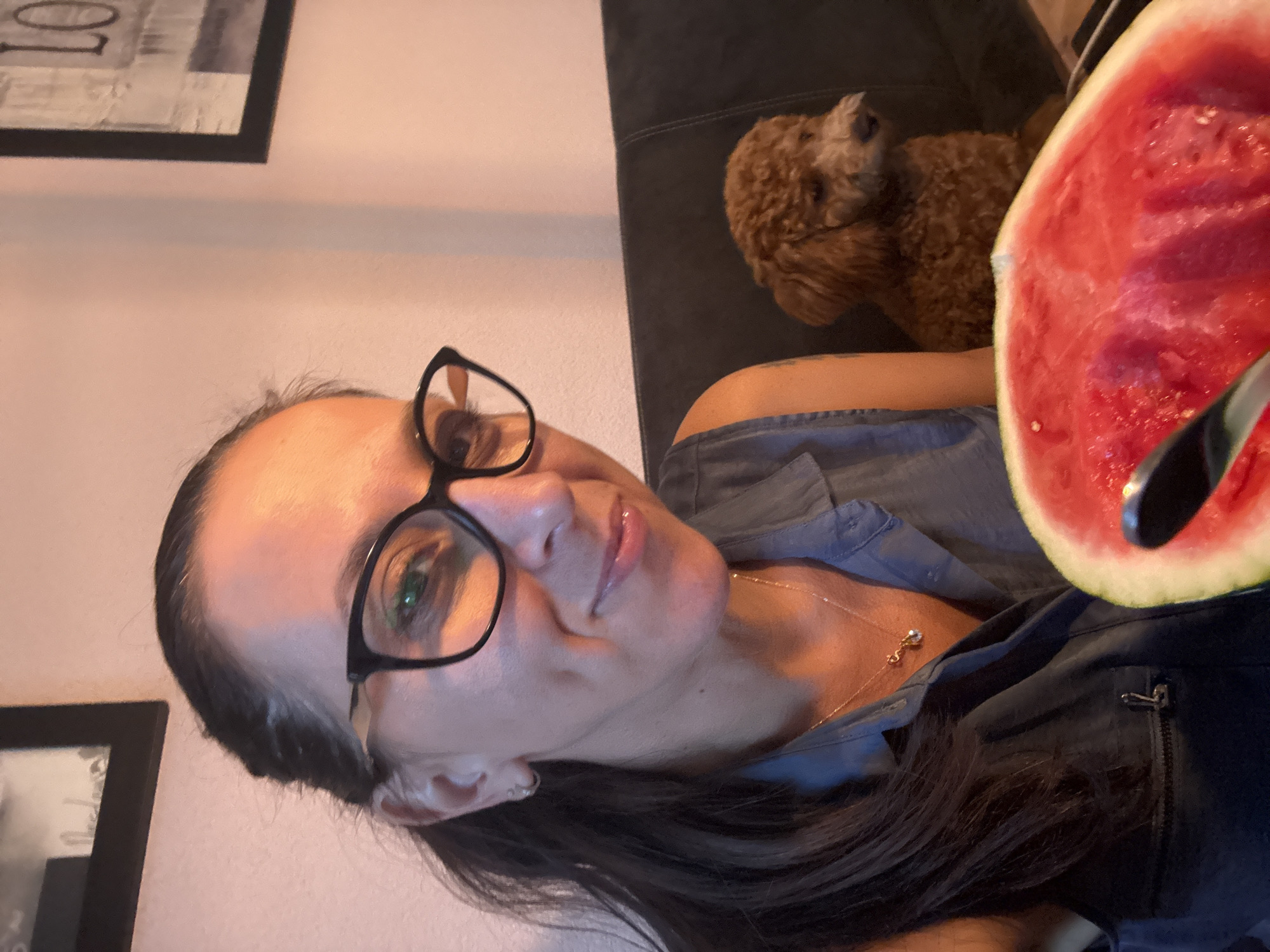 In an indoor setting, a woman with dark hair pulled back in a ponytail, wearing black-rimmed glasses and a blue shirt, smiles as she holds a slice of watermelon. She has dimples and is wearing a delicate necklace. A small, brown poodle sits on a dark couch behind her. The room's wall is a pale pink. Two framed pictures hang on the wall, one on the left and one on the right. The image does not provide any information about the city of Marum, Netherlands.