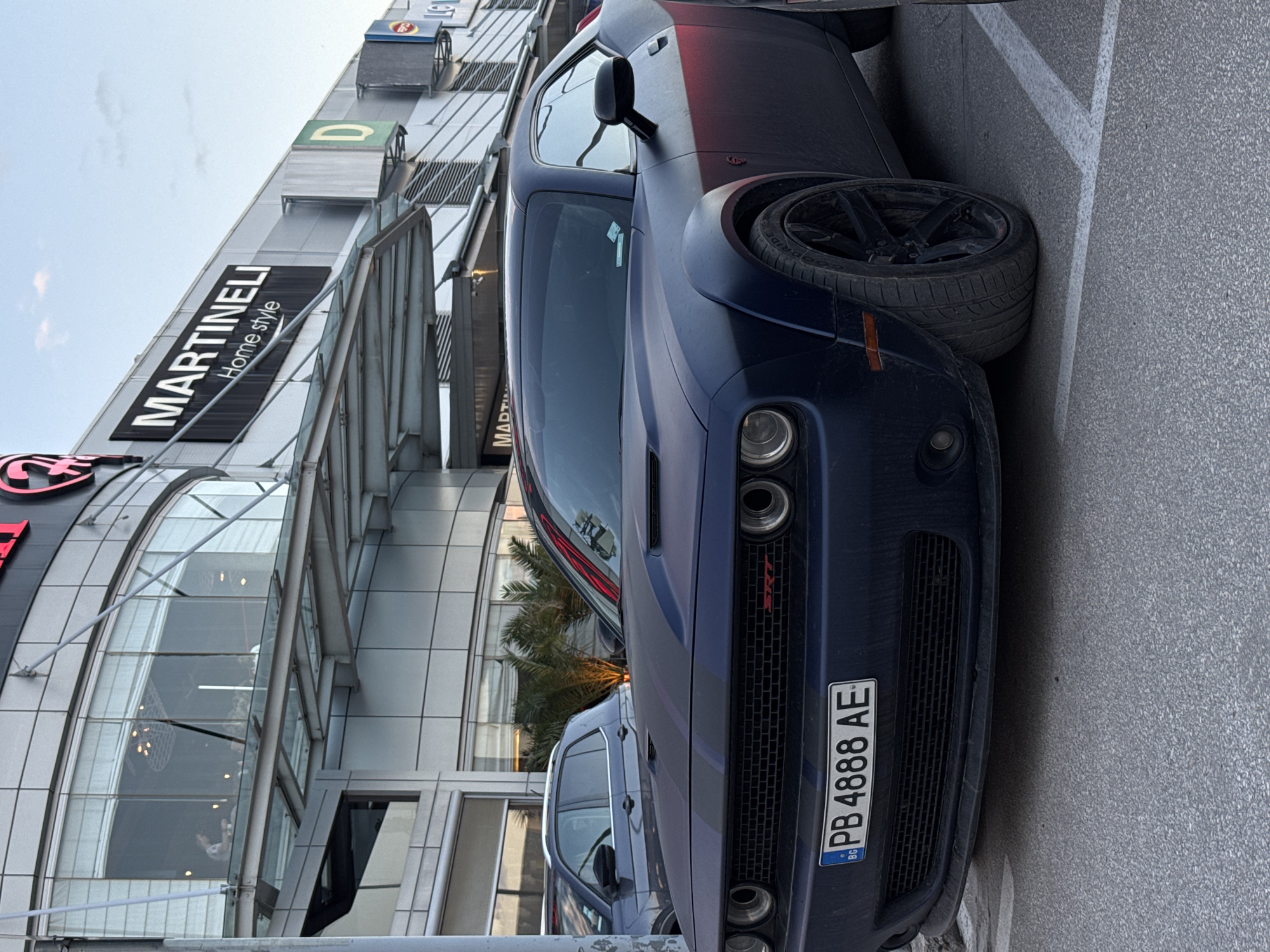 A ground-level, medium-shot depicts a blue Dodge Challenger parked at a sharp angle near a modern, multi-story building, identified by a "MARTINELLI Home style" sign. The vehicle's license plate, "PB 4888 AE", indicates registration in Bulgaria. The building features a glass facade with steel supports and reflects the sky and a palm tree. Part of another vehicle is visible behind the Dodge Challenger. The foreground shows an asphalt surface. The location is plausibly in the city of Plovdiv, Bulgaria, based on the license plate information.