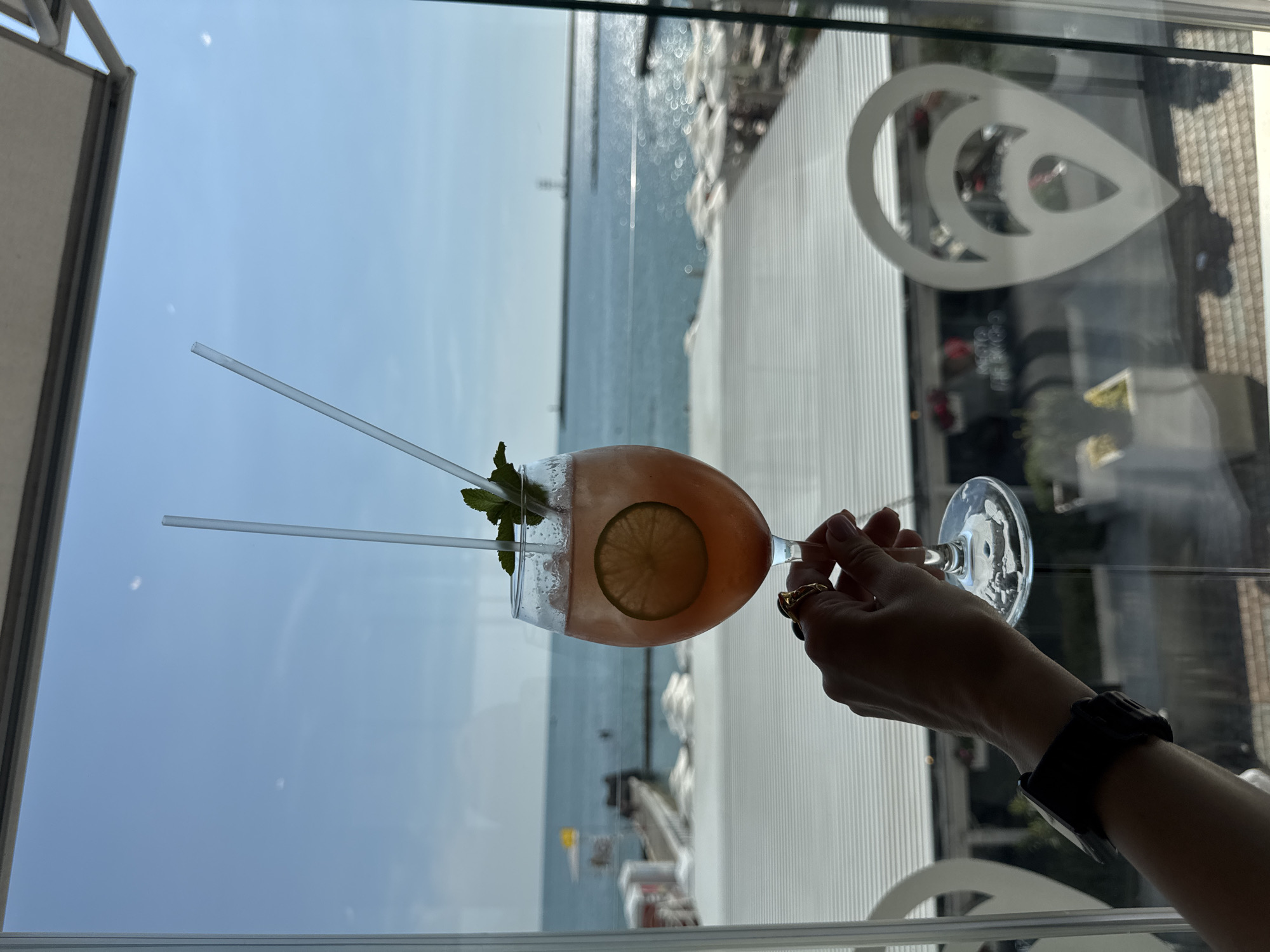 A hand, adorned with rings and a black watch, holds a stemmed glass containing a pink cocktail with two straws and a lime slice, against a backdrop of a bright, clear sky and the blue sea. The scene appears to be from a location in Sochi, Russia, possibly a seaside cafe or restaurant, as suggested by the presence of beach umbrellas and a pier in the distance. The glass is positioned in the foreground, obscuring a portion of the view of the outdoor seating area and the resort's reflective window.