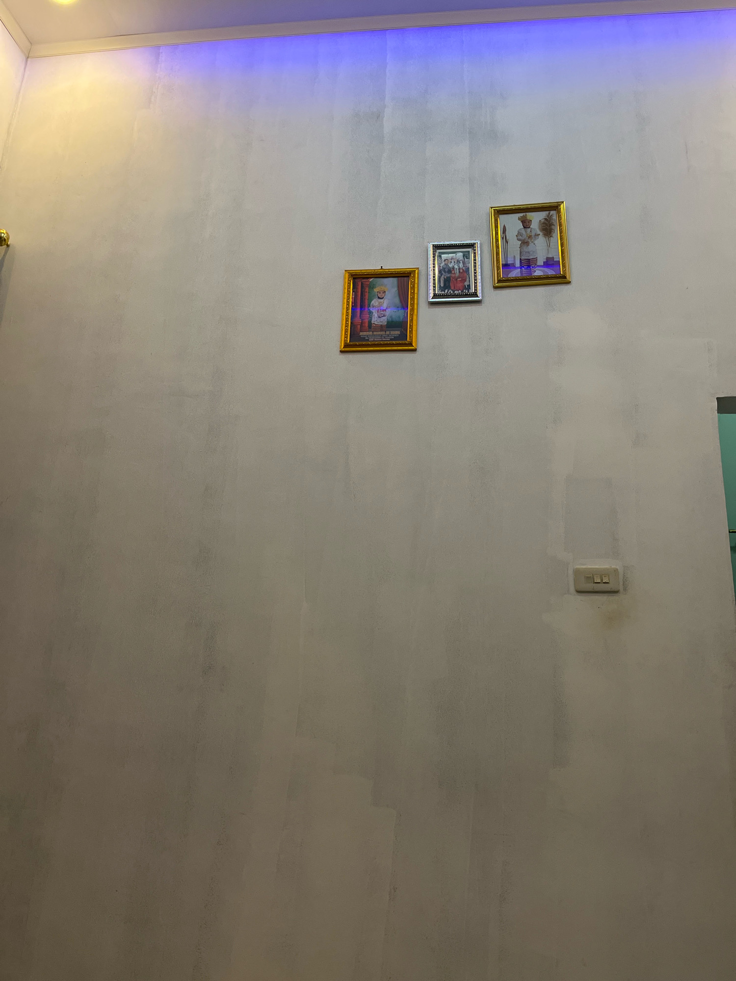 The image depicts a section of a wall in a room, likely in Kebumen, Indonesia. The wall is painted a light, neutral color and appears textured, with some subtle variations in shade. Three framed pictures are mounted on the wall. The leftmost picture has a gold frame and contains an image of a person in traditional attire. To its right, a smaller silver-framed picture shows a group of people, possibly a family, posing for a formal portrait. The third picture, also with a gold frame, features a person, again in traditional attire. Above the pictures, a blue LED light strip is visible along the ceiling edge, casting a soft glow. On the lower right side of the wall, a light-colored electrical switch plate with three switches is mounted. The overall impression is of a domestic interior space.