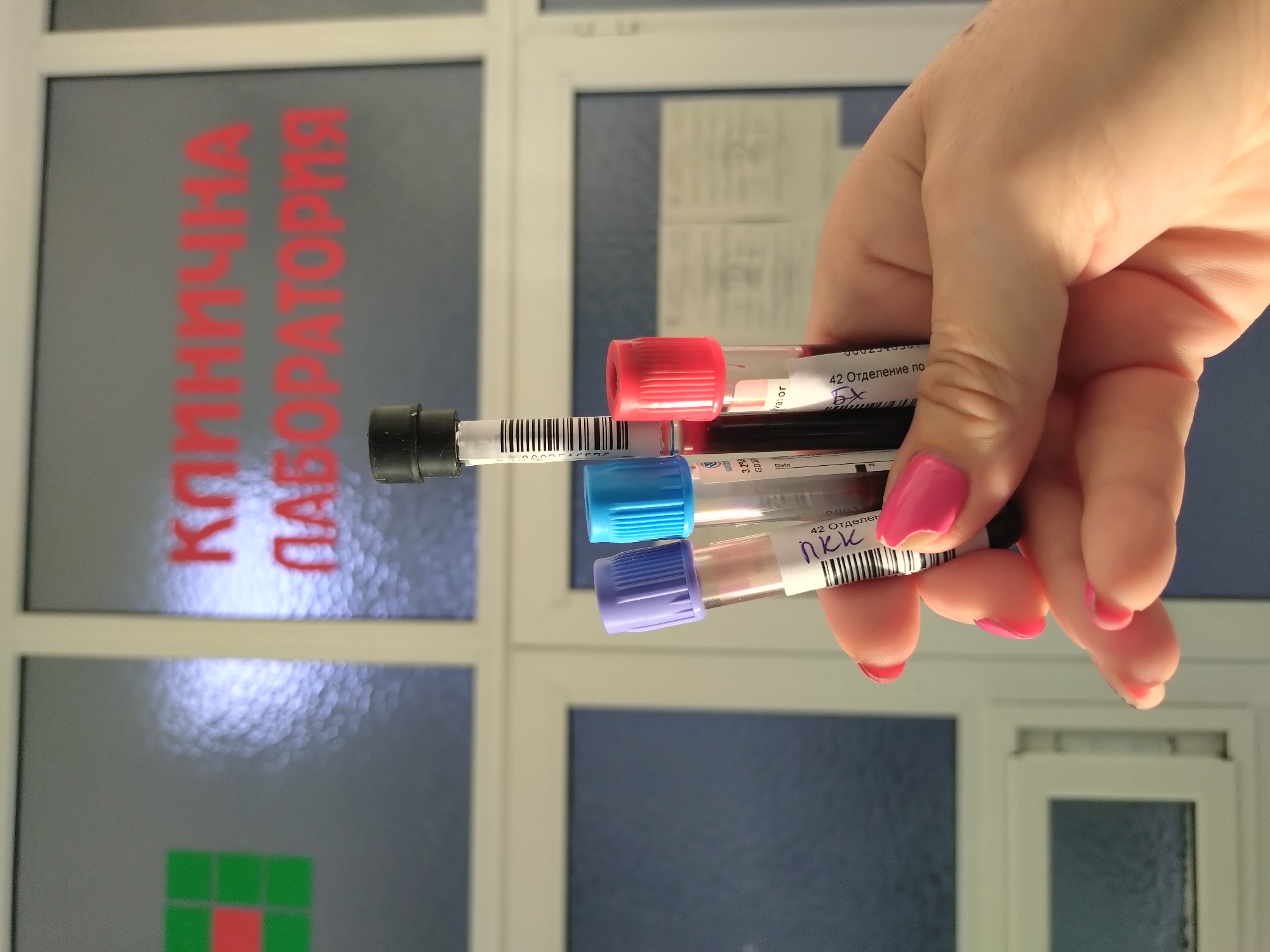 A hand with pink nail polish holds four blood collection tubes with different colored tops (red, black, blue, and lavender) in front of a window. Red lettering, "КЛИНИЧНА ЛАБОРАТОРИЯ", is visible on the window behind the tubes, suggesting a clinical laboratory. The label "42 Отделение по" is visible on one of the tubes. The location is Stara Zagora, Bulgaria, inferred from the Cyrillic text.