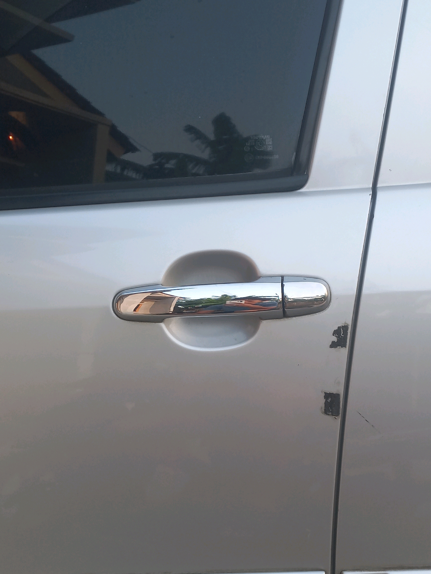 The image displays a close-up, eye-level view of the driver's side door of a silver car. A chrome door handle is prominently featured, reflecting a building and foliage. The door appears to have some minor damage, with chipped paint visible near the edge where the door meets the car's frame. The background is blurred, but the scene suggests an outdoor setting, possibly in Gambiranom, Indonesia, given the context. The lighting indicates it is daytime, with soft shadows cast on the car's surface.