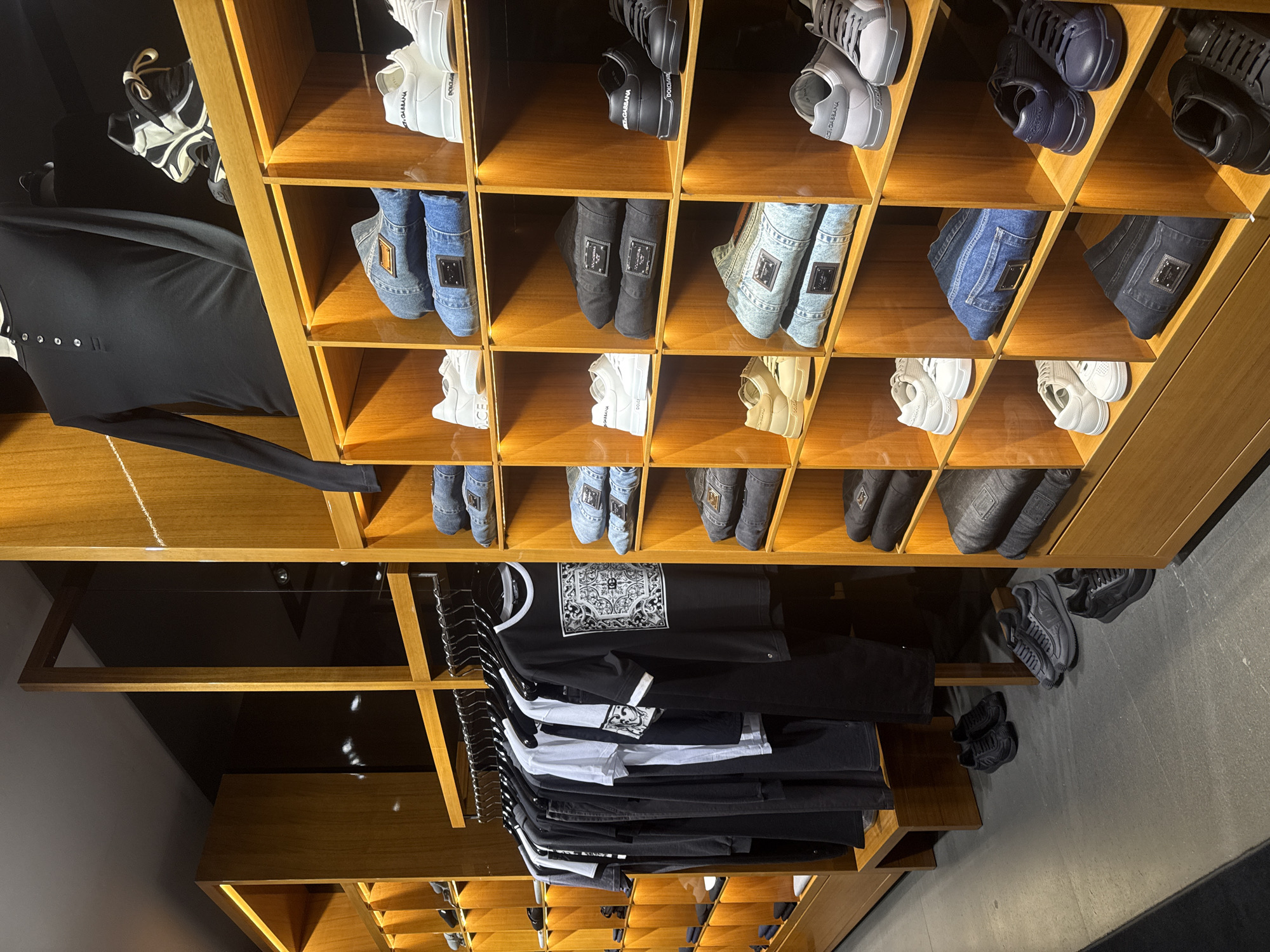 The image depicts an indoor retail setting in what appears to be a clothing store or high-end boutique. The focus is on a display unit constructed of wooden shelving, arranged in a grid of cubbies or compartments. The shelves are stocked with various items, primarily shoes and garments. The shoes are diverse in style, ranging from sneakers to slip-on styles, and are predominantly black, white, and blue denim. Some shoes have visible brand labels. On the clothing shelves, a rack holds a selection of t-shirts and long-sleeved shirts, mostly in black and white, some with graphic designs. The clothing appears to be neatly stacked and hung. The environment is well-lit, with the wooden display unit appearing clean and orderly. The presence of the clothing and shoes suggests a retail setting, potentially specializing in designer or high-end apparel. No people are visible in the frame, and based on the limited information, it is not possible to confirm if the setting is in Yekaterinburg, Russia, without additional data or context.