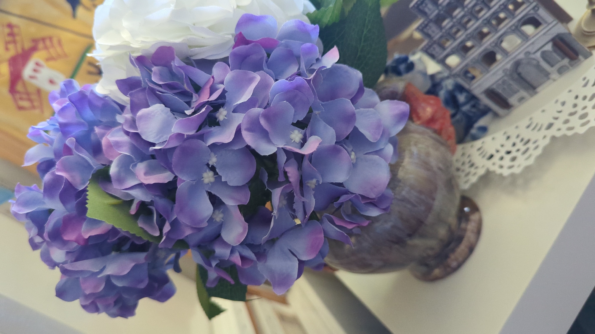 The image shows a close-up of a bouquet of purple hydrangeas in a decorative vase. To the right of the hydrangeas, a white flower is partially visible. The vase has a marbled pattern and is placed on a light-colored surface, possibly a table or shelf. In the background, a colorful tapestry or painting is hanging on a wall. Partially visible on the surface in front of the vase is a small, gray, architectural model that resembles a coliseum, and a white lace trim. The lighting suggests an indoor setting, possibly a home or a shop. The image is cropped to focus on the floral arrangement and surrounding objects, with the background out of focus. The location in Kløfta, Norway is not specified or evident from the image itself.