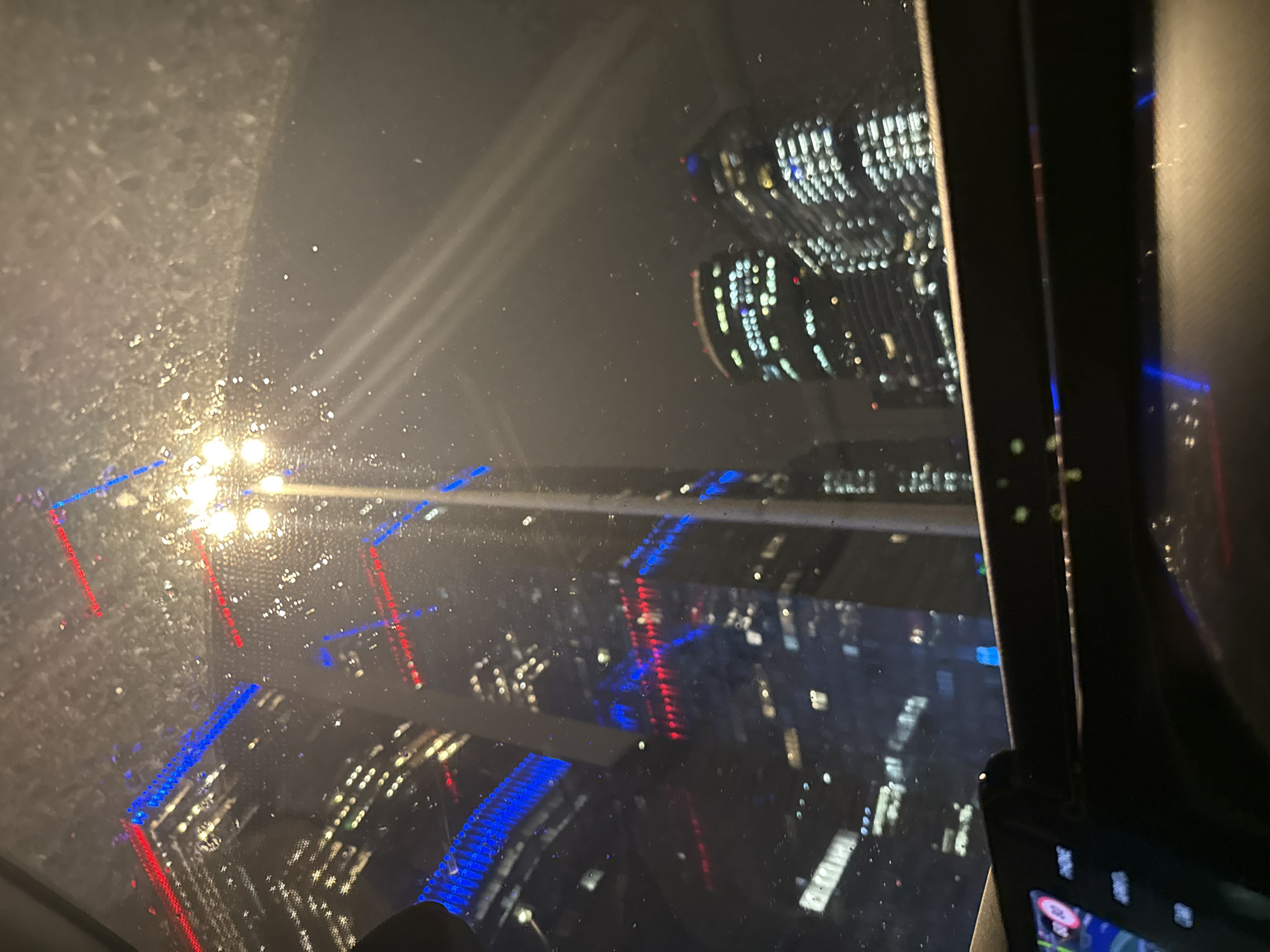 This is a nighttime, low-angle shot from inside a vehicle, looking through a wet window at a city skyline. The foreground shows the dashboard of the car, with a display screen on the left. The midground features the city skyline with several tall buildings illuminated by numerous lights. Some buildings have streaks of blue and red lights, likely decorative or functional architectural lighting. The wet window causes reflections and distortions of the lights and buildings. A streetlamp is visible in the upper right quadrant of the image. The overall atmosphere is dark, moody, and urban, possibly during or after rain.