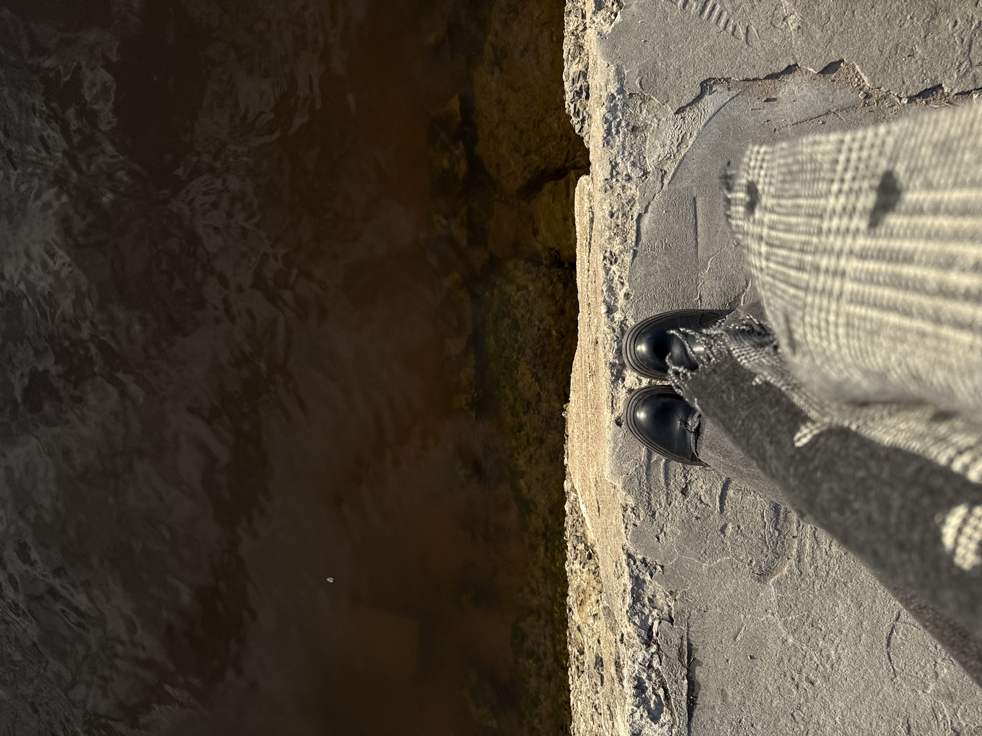 A high-angle shot shows a person standing on a weathered concrete edge next to dark water. The person's legs are visible, clad in plaid pants and black boots. The edge appears to be part of a canal or riverbank in Saint Petersburg, Russia, based on the architectural style of the concrete. The water is murky, and some rocks and possible algae growth can be seen beneath the surface near the edge. The concrete shows signs of age and wear, with cracks and rough textures. A footprint is visible on the concrete.
