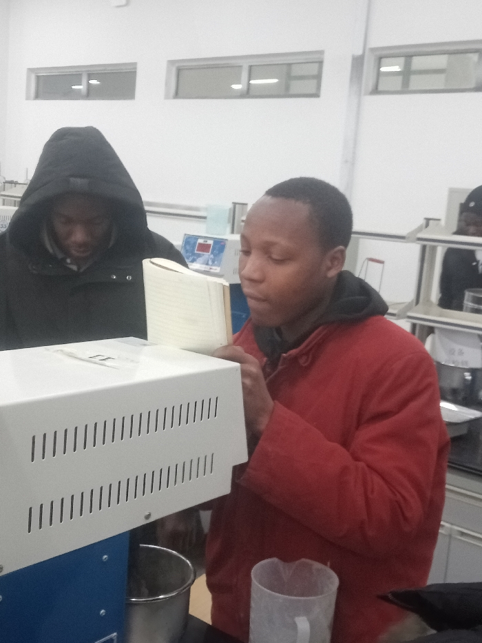 Two men are in a brightly lit laboratory setting. The man on the right is wearing a red jacket. He is focused on operating a piece of machinery. A notebook is visible next to the machine. The other man, wearing a black hooded jacket, observes the process. Laboratory equipment and countertops line the background, with windows offering natural light.