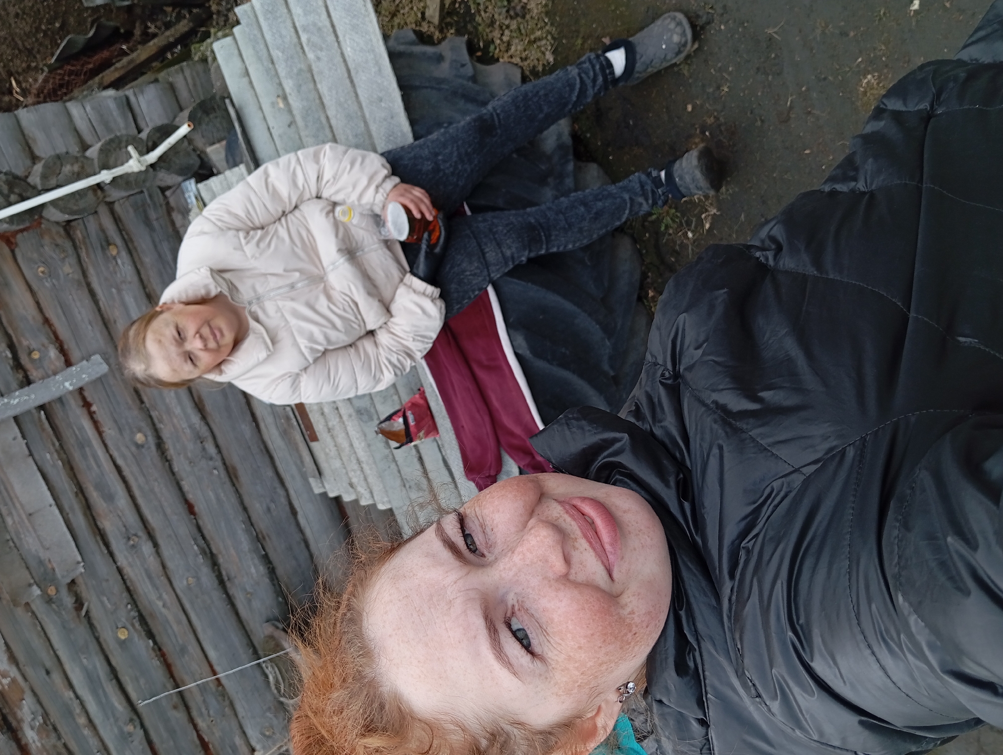 Two women are visible in a selfie-style photograph taken in Yurgamysh, Russia. One woman with red hair and freckles is taking the picture, wearing a black padded jacket. The other woman is further away and appears to be sitting or lying down. She is wearing a light beige puffy jacket, dark-colored pants, and shoes. She is holding a cup. They are on a surface constructed of weathered wood and metal, with an area of bare ground adjacent to them. Other objects such as a red-colored garment are present in the scene.