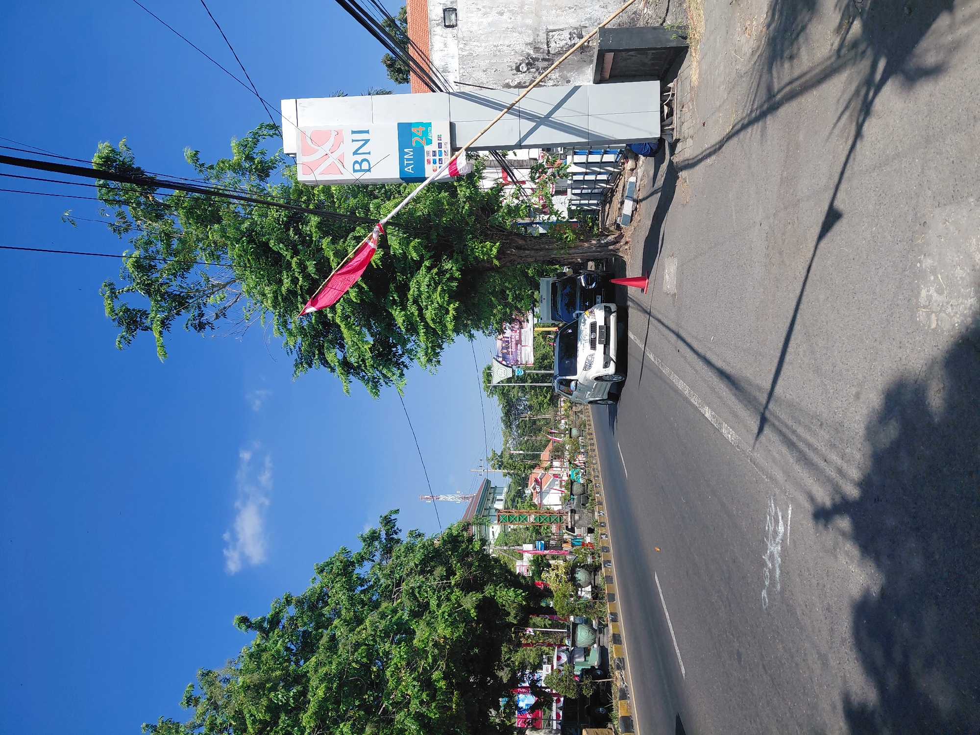 The image depicts a street scene in Semampir, Indonesia, on a clear, sunny day. A paved road runs from the left foreground towards the background, bordered by trees and vegetation on the left and a sidewalk and a building on the right. A white car and a black car are parked on the side of the road, near a red traffic cone. A large sign for BNI (Bank Negara Indonesia) with an ATM 24 Jam advertisement stands prominently on the right side of the image. A red and white flag is attached to a pole near the BNI sign. A person in blue is visible kneeling near the base of the BNI sign. Power lines are strung across the upper portion of the image, casting shadows on the road and sidewalk. Buildings and more trees are visible in the background, along with a cell tower. The overall impression is of a typical urban street environment.