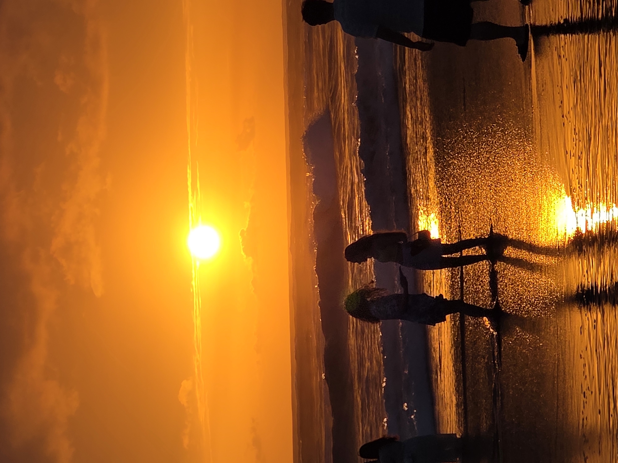 This is a vertical, full-shot, eye-level photograph of a beach at sunset in North Kuta, Indonesia. The sun is a bright, white orb situated slightly to the left of the horizontal center of the frame, positioned high in the sky. The sky is a gradient of intense orange and yellow, with some wispy clouds visible. The sun's light casts a warm, golden hue over the entire scene, illuminating the ocean waves and the wet sand.
In the foreground, two women, silhouetted against the bright sky, stand close to each other on the wet sand. They appear to be interacting, with one woman gesturing with her hands. Both have long, dark hair. To the left of these two women, a third person, also silhouetted, stands partially in the water, facing away from the camera. To the far right of the frame, a man stands with his back to the camera, his silhouette dark against the vibrant sunset. He is wearing a t-shirt and shorts, and his feet are in the shallow water.
The ocean is visible in the middle ground, with moderate waves breaking and creating white foam. The wet sand in the foreground reflects the colors of the sunset, creating a shimmering, golden surface. The overall impression is one of a serene and beautiful evening at the beach.