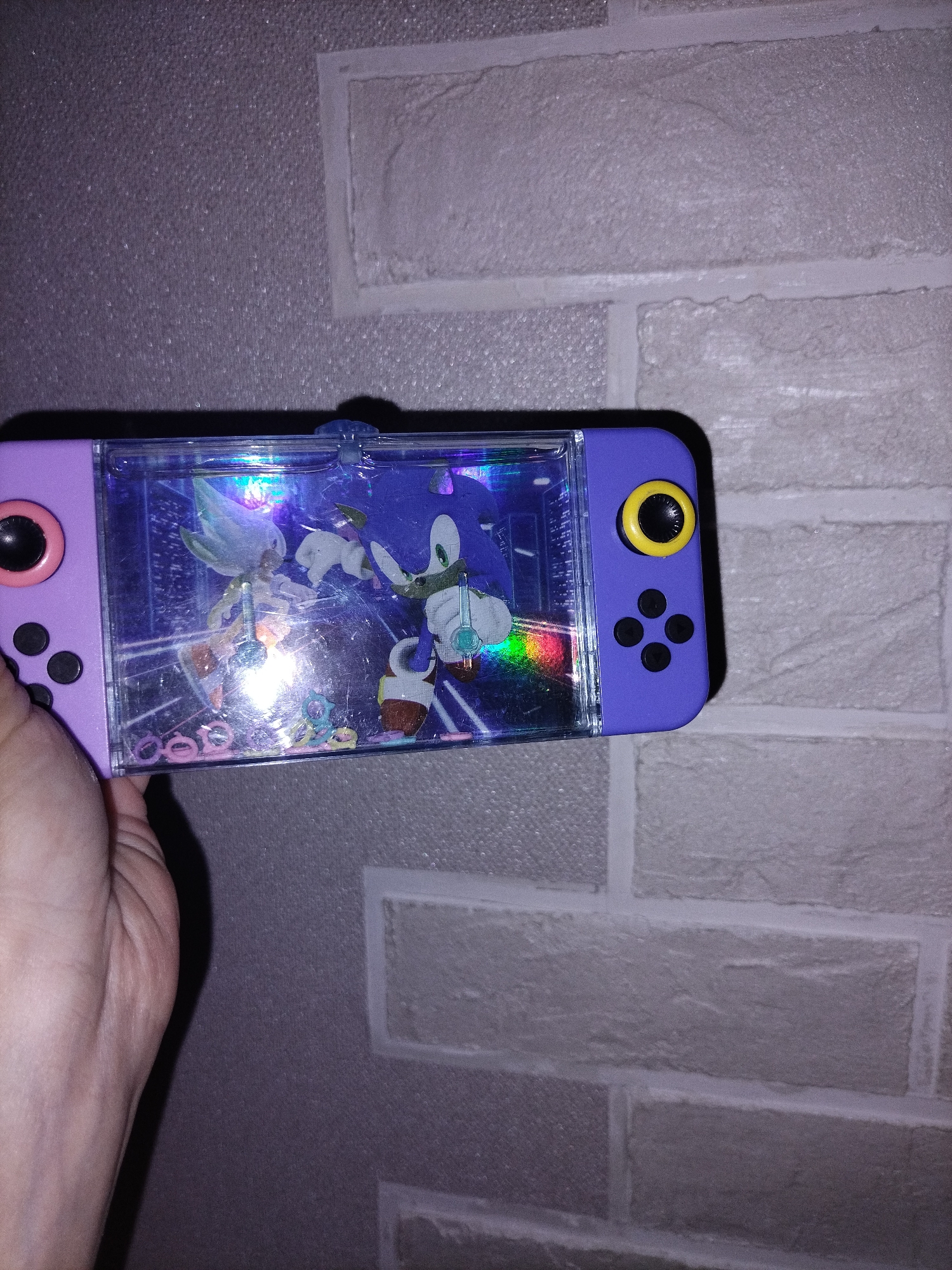 A hand holds a purple, handheld water game or liquid-filled toy case against a light-colored, textured brick wall. The toy is oriented horizontally. Its casing is a vibrant purple, featuring a joystick with a pink ring and black center on the left, and a joystick with a bright yellow ring and black center on the right. Below each joystick are four small, black circular buttons arranged in a diamond pattern.
The central part of the toy is a clear, rectangular chamber filled with liquid, within which figures resembling Sonic the Hedgehog (blue) and Silver the Hedgehog (white/silver) are positioned. Small, colorful floating objects, possibly rings or charms, are also visible within the liquid at the bottom of the chamber. Significant glare and reflections, including a rainbow-like shimmer, are present on the clear plastic surface, partially obscuring the figures inside.
The background wall displays a distinct brick pattern, rendered in shades of light gray or off-white, suggesting an indoor setting. The lighting appears to be artificial, casting some shadows and highlighting the reflective surfaces of the toy. No legible text is visible in the image.