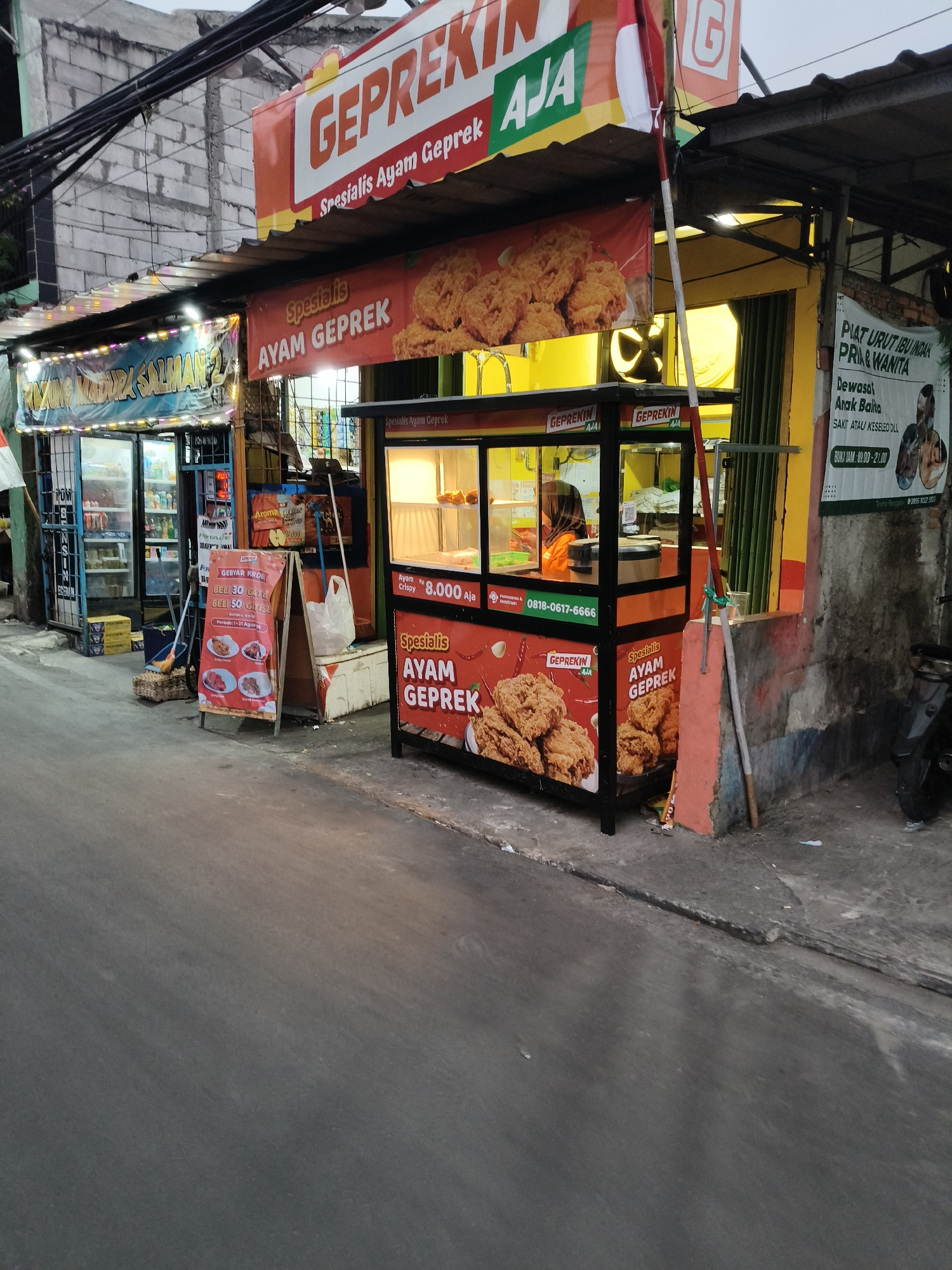 The image depicts a street-level view of several food stalls in South Tangerang, Indonesia. The primary focus is on a "GEPREK KIN AJA" stall, which specializes in "Ayam Geprek" (smashed fried chicken). This stall features a brightly colored facade with images of fried chicken and promotional banners. A person wearing a hijab and an orange shirt is visible inside the stall. Next to it, another stall is partially visible, displaying various drinks and snacks. A prominent red A-frame sign in front of one of the stalls advertises a promotion: "BELI 30 GRATIS BELI 50 GRATIS." Further down the street, there are other structures and a motorcycle parked on the right side. The environment suggests an urban street with a modest level of commercial activity, likely in the evening or early morning given the lighting conditions. The buildings in the background appear to be constructed from brick and concrete.