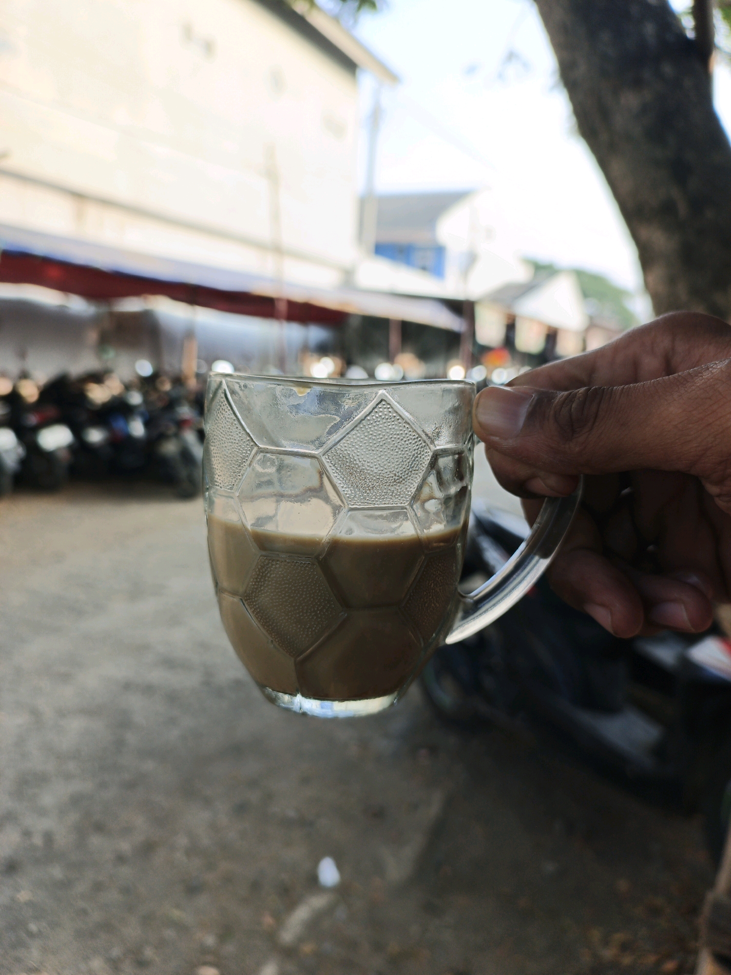 A hand is holding a clear glass mug filled with a light brown liquid, likely coffee or tea, in an outdoor urban setting. The mug has a hexagonal pattern etched into its glass. In the background, there are several motorcycles parked in rows. The environment appears to be a street or an open area with buildings and trees visible. The lighting suggests it is daytime. The location is identified as Krimun, Indonesia.
