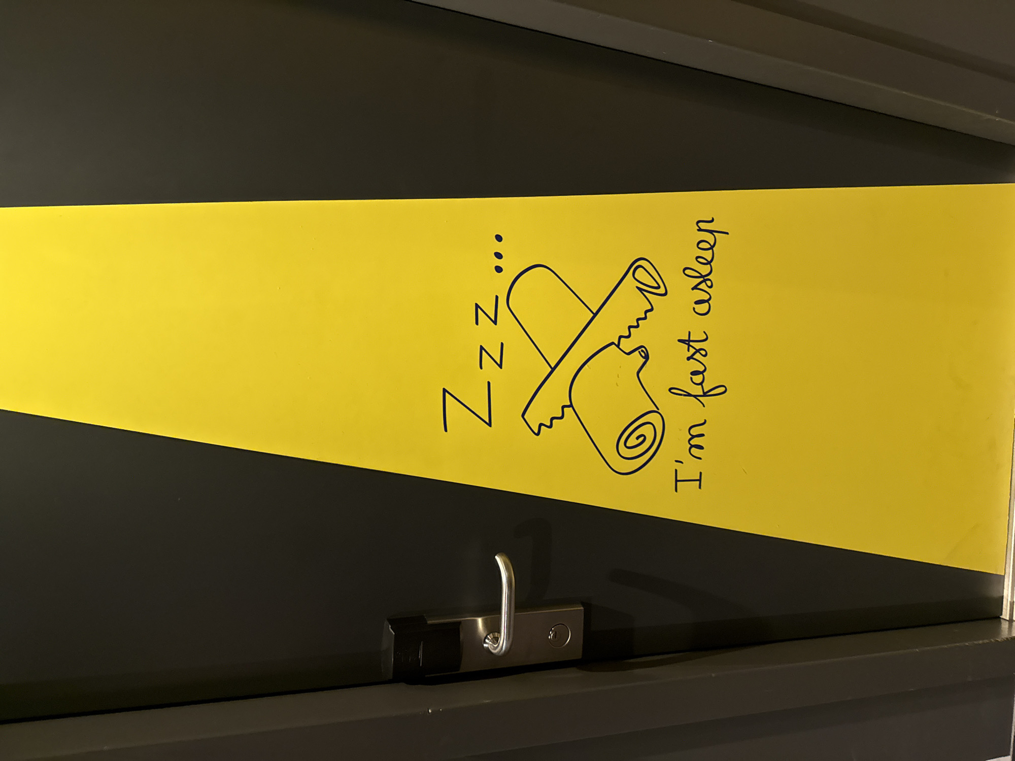 A close-up, eye-level shot shows a black door with a yellow diagonal stripe running down the right side. A silver door handle and a black electronic lock are visible on the left side of the door. On the yellow stripe, a black line drawing depicts a rolled-up sleeping bag with a jagged edge, and the text "Zzz ..." and "I'm fast asleep" are written in black cursive. The image was taken in Stein, Netherlands, at the Kasteelpark.