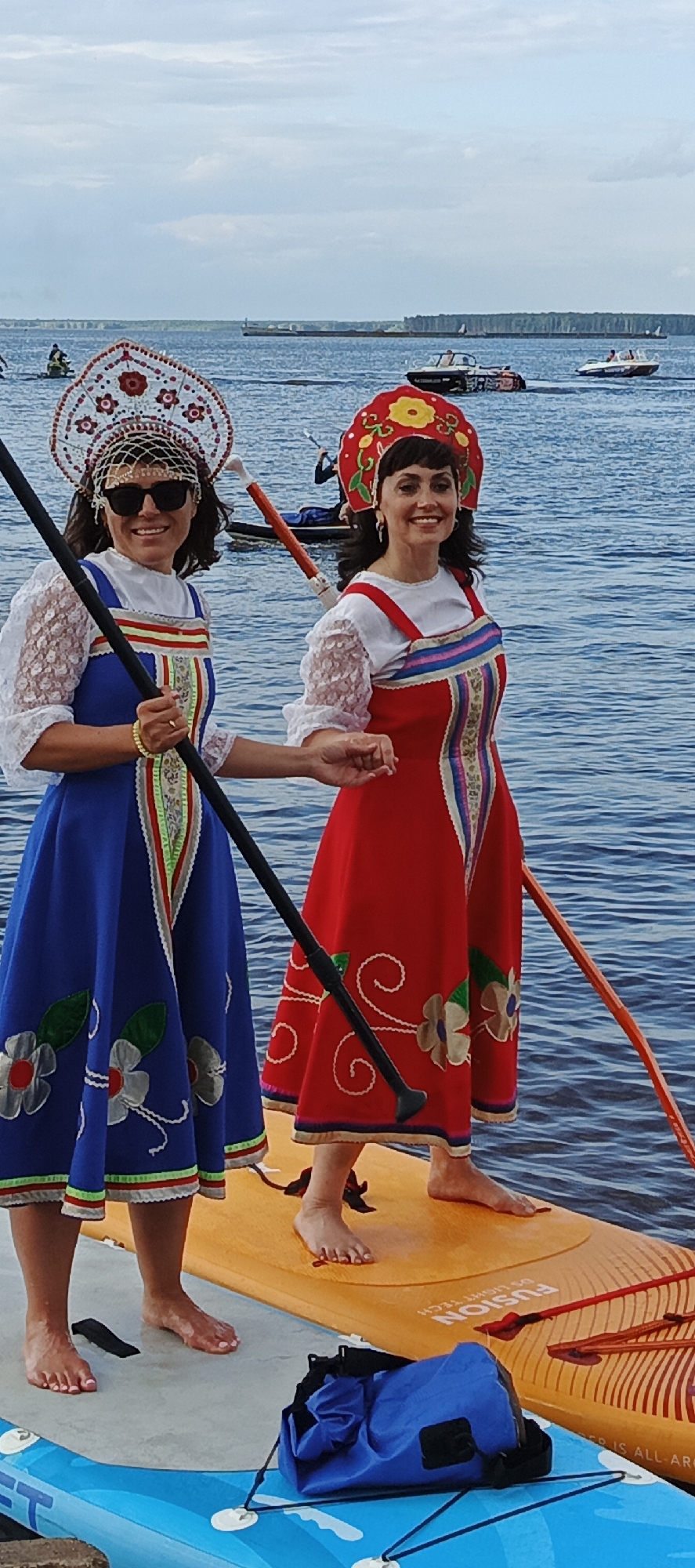 Two women in traditional Russian attire, including kokoshnik headdresses, stand on paddleboards in the Kama River in Chaikovsky, Russia. The woman on the left wears a blue dress and the woman on the right wears a red dress. Both are holding paddles. The river is calm, with a few boats visible in the background. The sky is overcast.