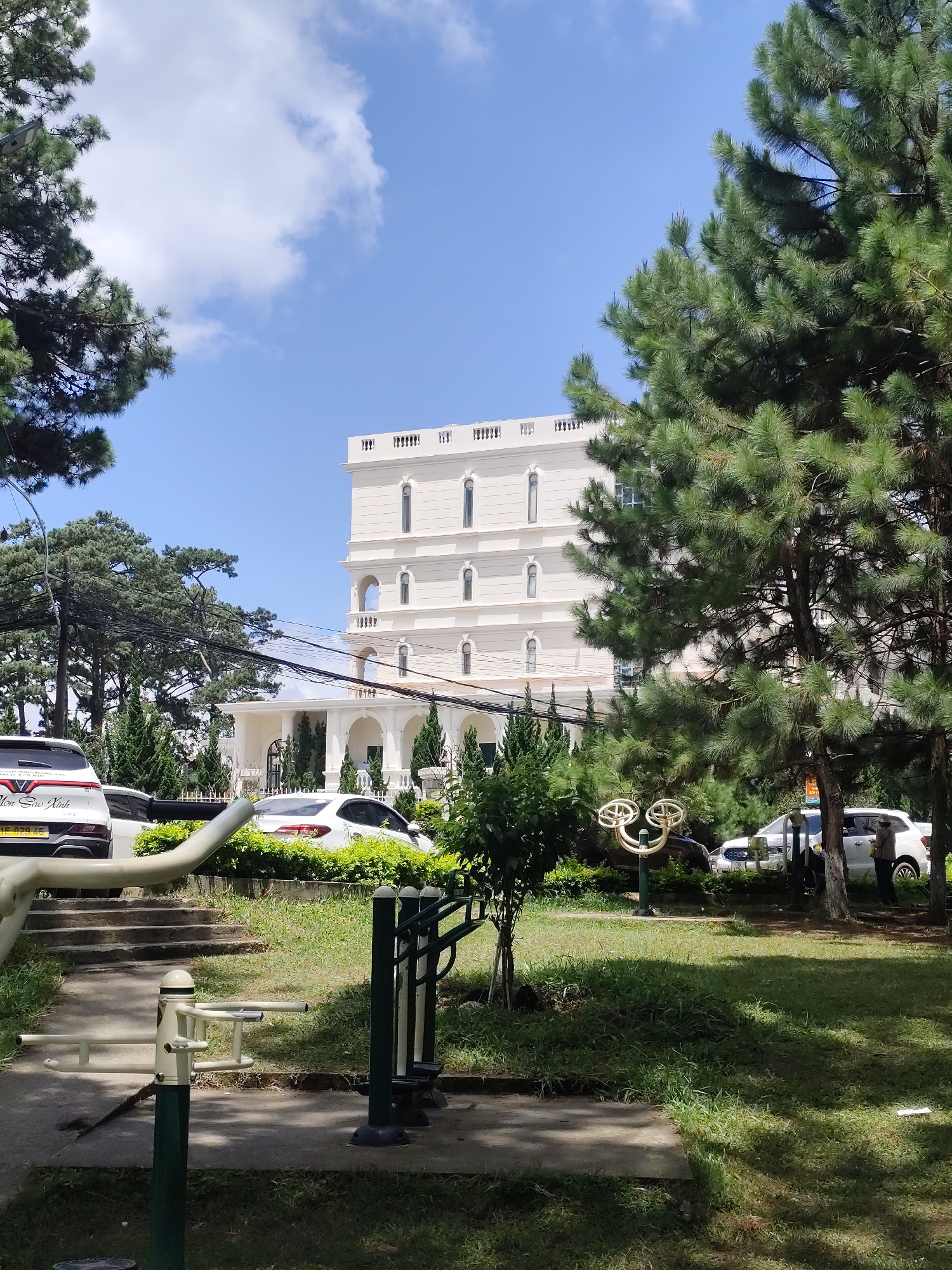 The image depicts an outdoor scene in Da Lat, Vietnam, with a focus on a white multi-story building in the background. The building has a classical architectural style with arched windows and decorative elements along the roofline. In the foreground, there are several pieces of outdoor exercise equipment, including what appears to be a seated leg press machine and a body twist machine. A paved pathway and grassy areas are visible, along with several parked cars, one of which is a white SUV with a license plate starting with "ME". Tall pine trees dominate the right side of the frame, with their branches casting shadows on the ground. The sky is blue with scattered white clouds, suggesting a sunny day. The overall impression is of a well-maintained public space or park adjacent to the building.