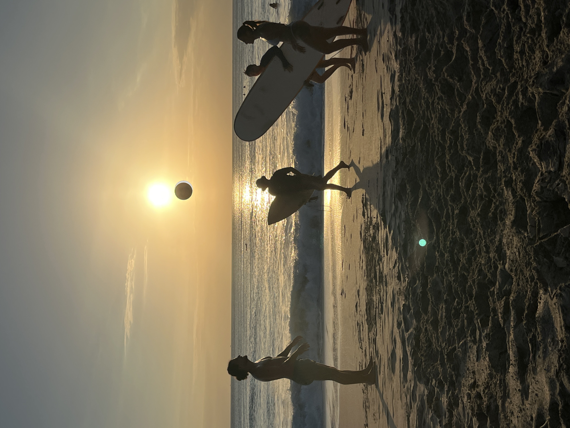 In Kuta Selatan, Indonesia, at sunset, four individuals are silhouetted on a sandy beach. The sun, a bright orb low in the sky, casts a golden light across the scene, illuminating the ocean waves and the wet sand. A dark, circular object, possibly a ball, is suspended in the air between the sun and the horizon.
To the left, a man stands facing the ocean, his body in profile, appearing to be in a relaxed stance. In the center, another individual, also silhouetted, walks away from the viewer, holding a surfboard. To the right, two people are seen from behind, carrying a large surfboard between them, also facing the ocean. The surfboards are all light-colored, contrasting with the dark silhouettes of the people. The foreground is dominated by the textured, dark sand of the beach, with a small, bright green object, possibly a piece of debris or a reflection, visible on the sand in the lower left quadrant. The ocean behind them is active with waves breaking. The sky transitions from a pale yellow near the horizon to a soft blue overhead, with a thin, wispy cloud visible in the upper left portion of the frame.